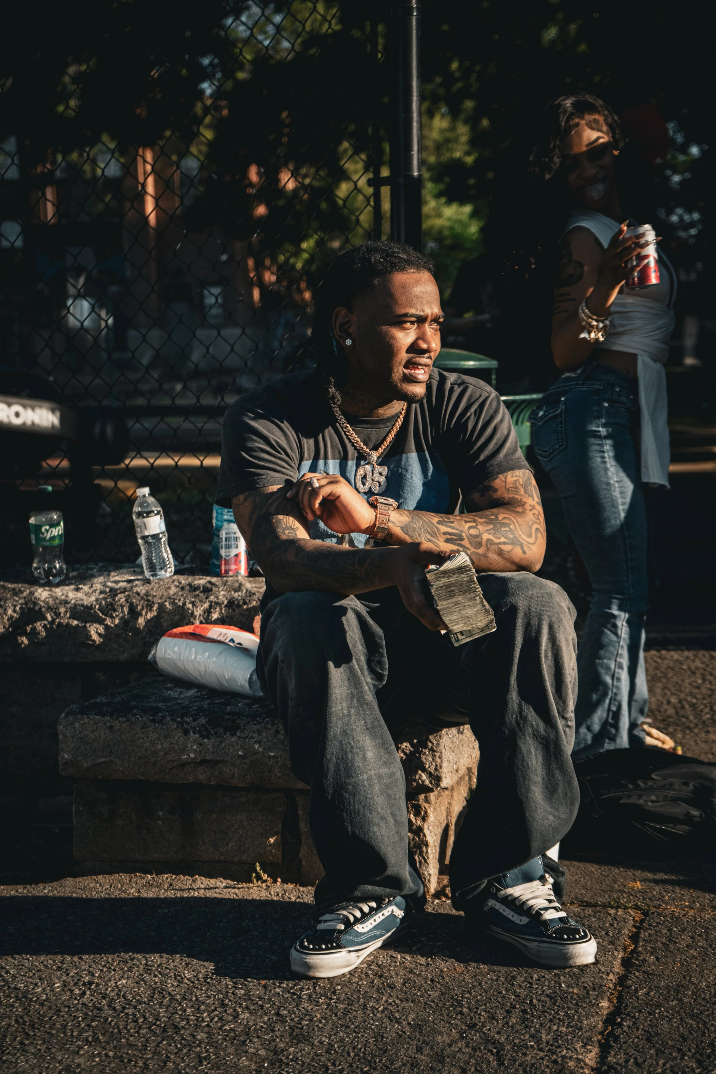 A young man with tattoos, wearing a black and gray t-shirt, dark jeans, and black and white sneakers, sitting on a stone bench outdoors. Behind him is a chain-link fence, with a woman standing next to him holding a soda can and smiling. Bottled drink