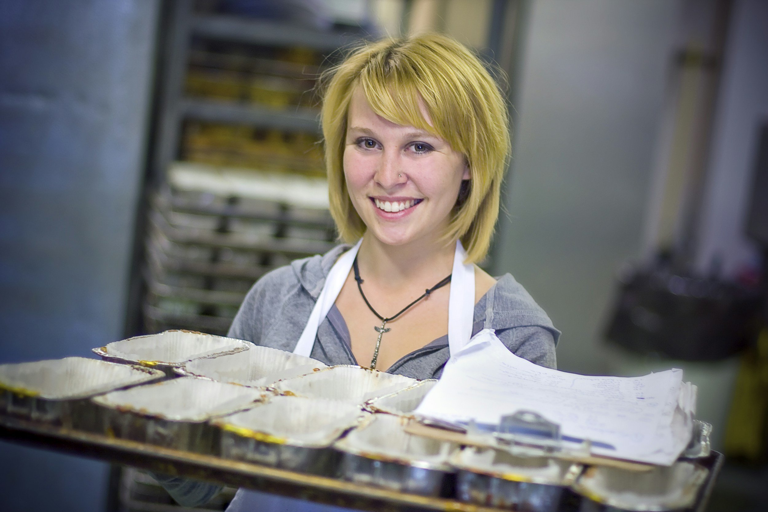 A smiling woman with short blonde hair wearing a gray hoodie, a white apron, and a necklace, standing in a kitchen near a tray of baked goods.