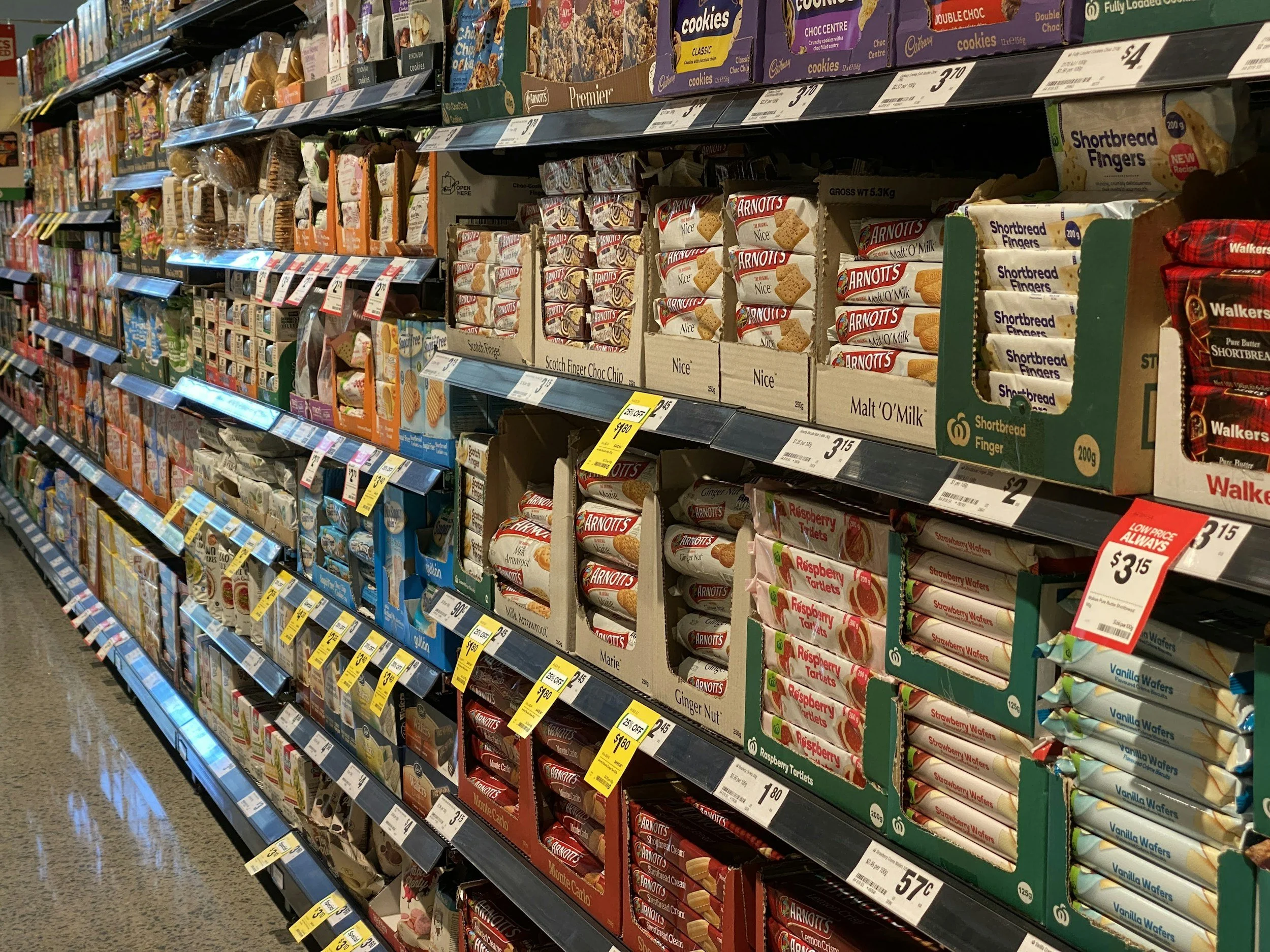 Aisle in a grocery store with shelves stocked with snack foods, including various brands of cookies, biscuits, and wafers, with price tags on the shelves.