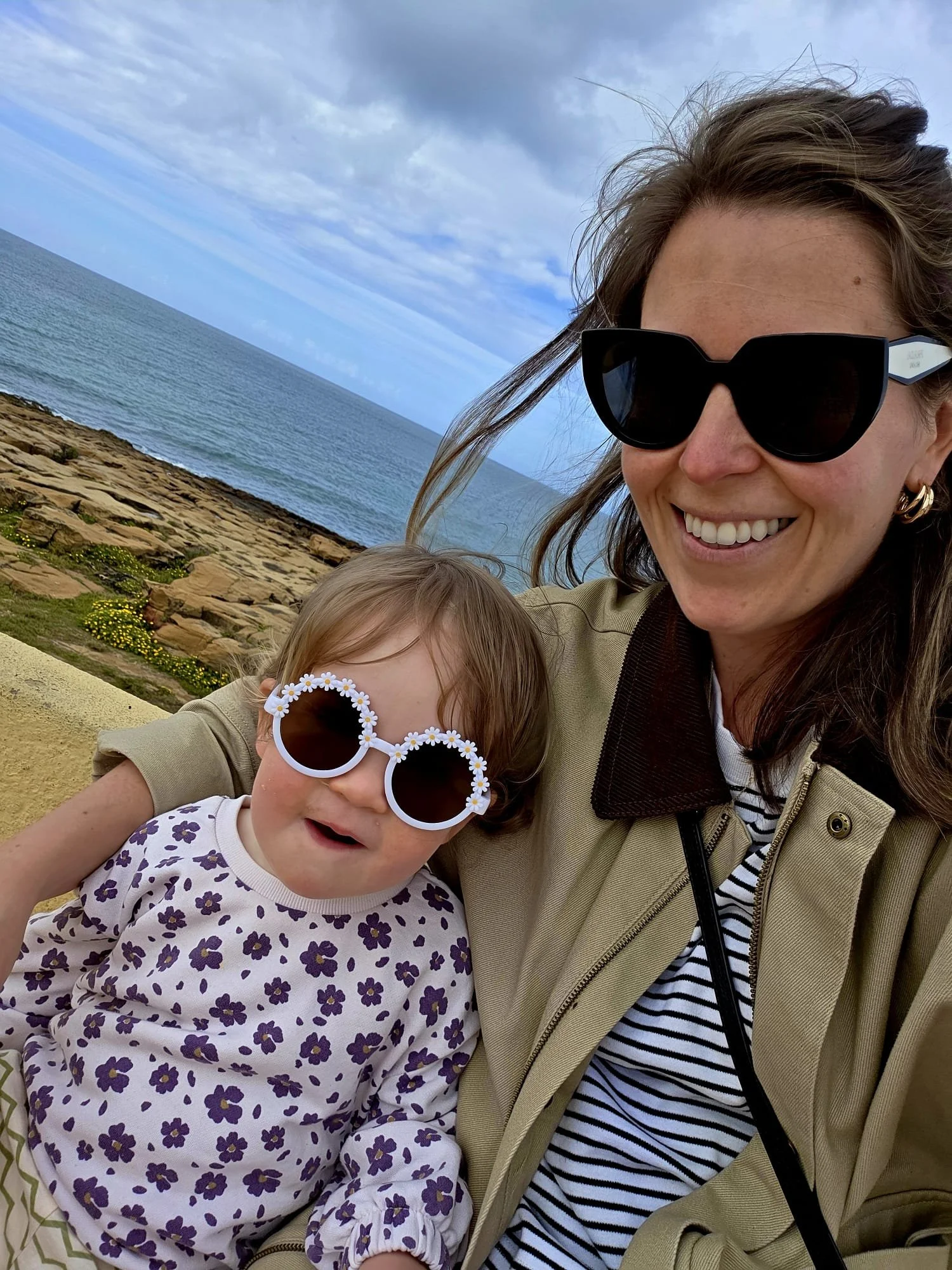 A woman and a young girl smiling at the camera while wearing sunglasses by the seaside. The woman wears black sunglasses and a beige jacket, and the girl wears white sunglasses decorated with daisies and a purple floral shirt.