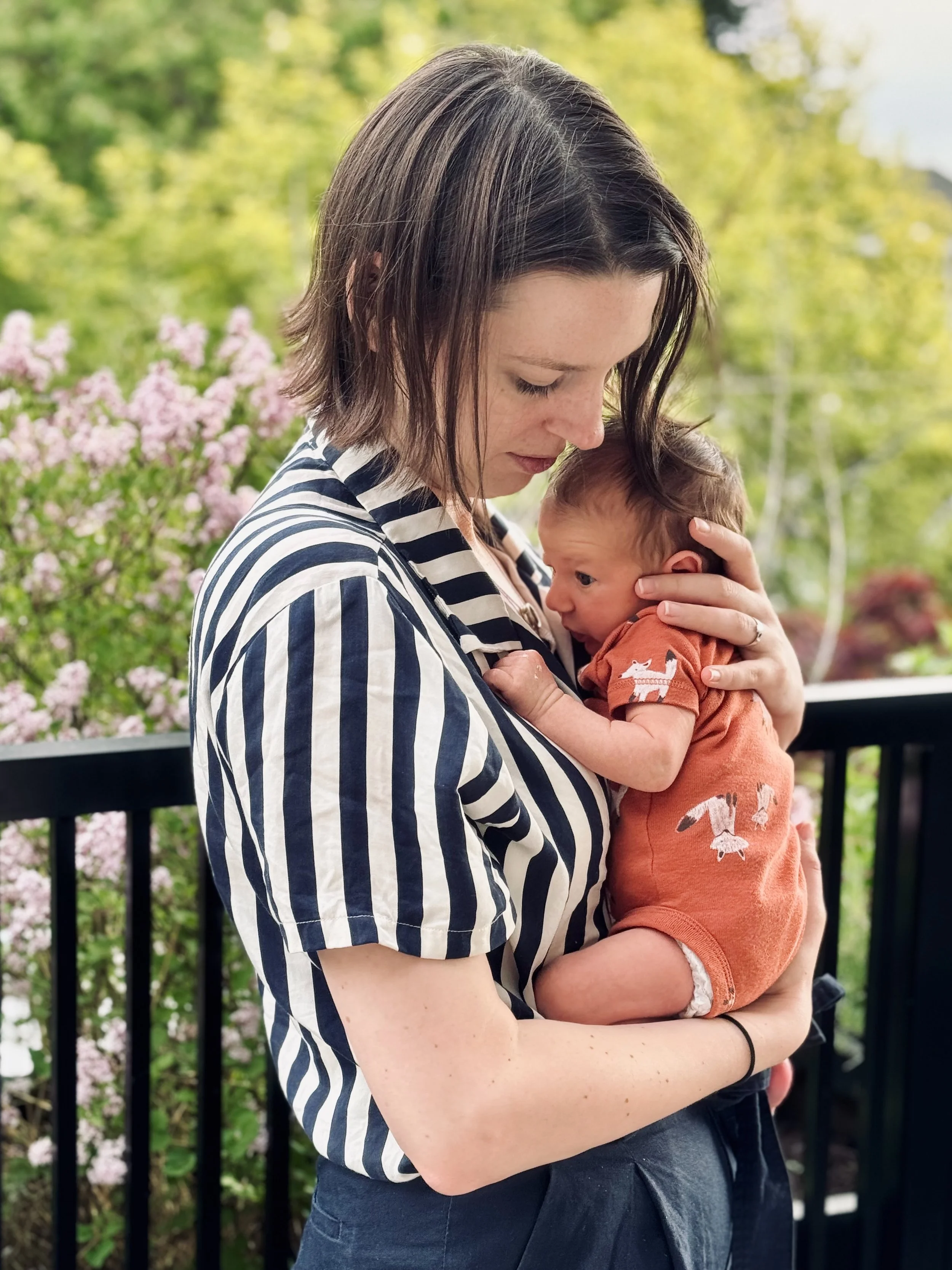 A woman with shoulder-length dark hair, wearing a black and white striped shirt, holding a small baby with short brown hair, dressed in an orange outfit with a bird pattern, outdoors near a black railing with pink flowering bushes and green trees in the background.