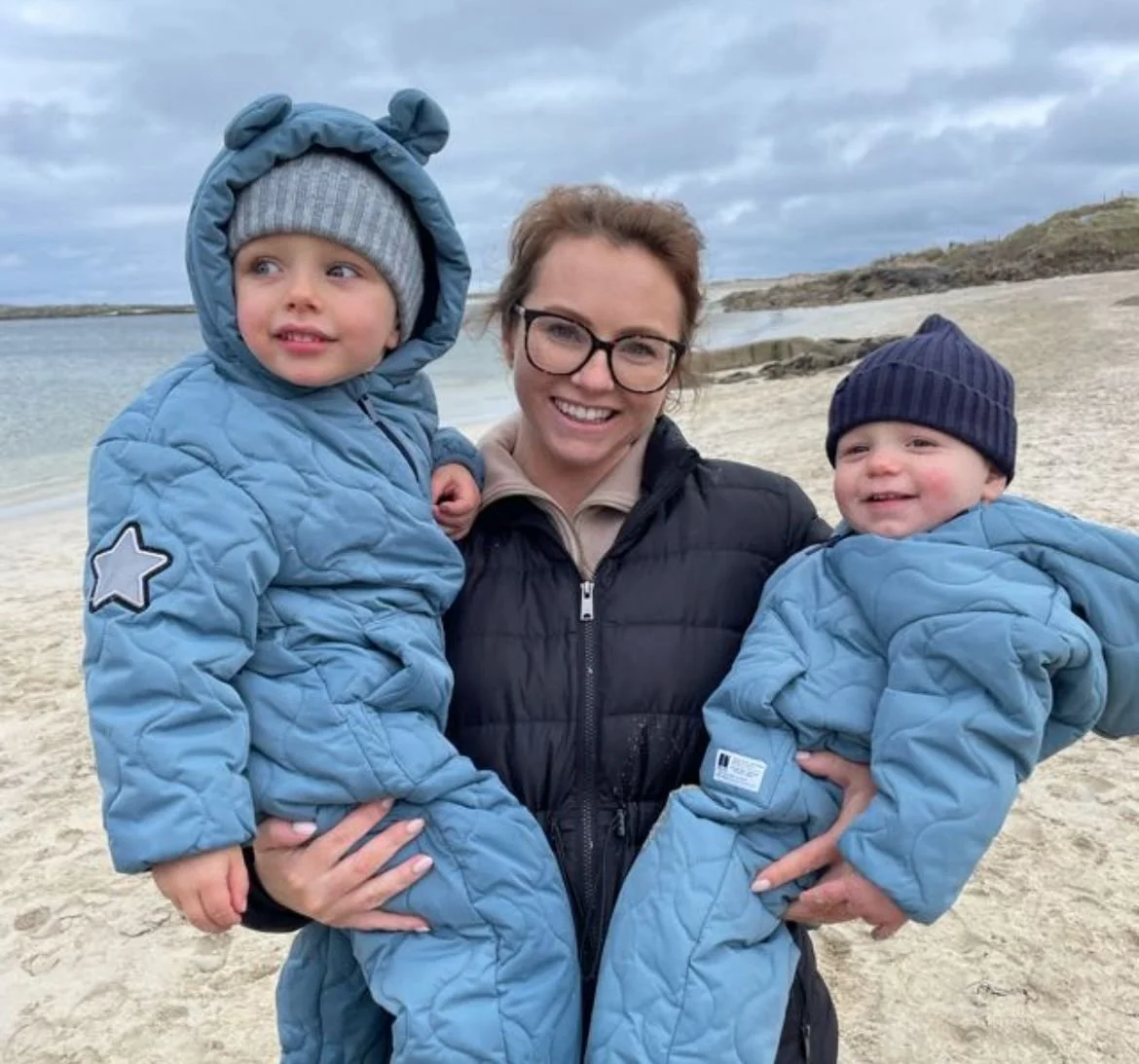 A woman holding two young children on a sandy beach with a cloudy sky in the background. All are dressed in blue winter jackets and hats, smiling at the camera.