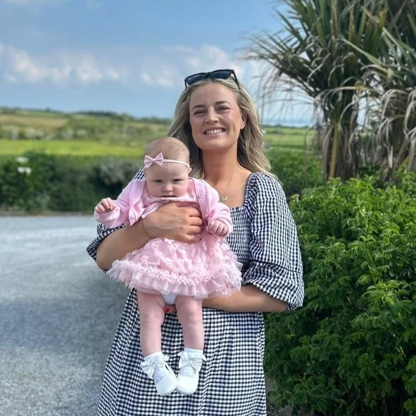 A smiling woman holding a baby girl dressed in pink outdoors near a body of water and green foliage.