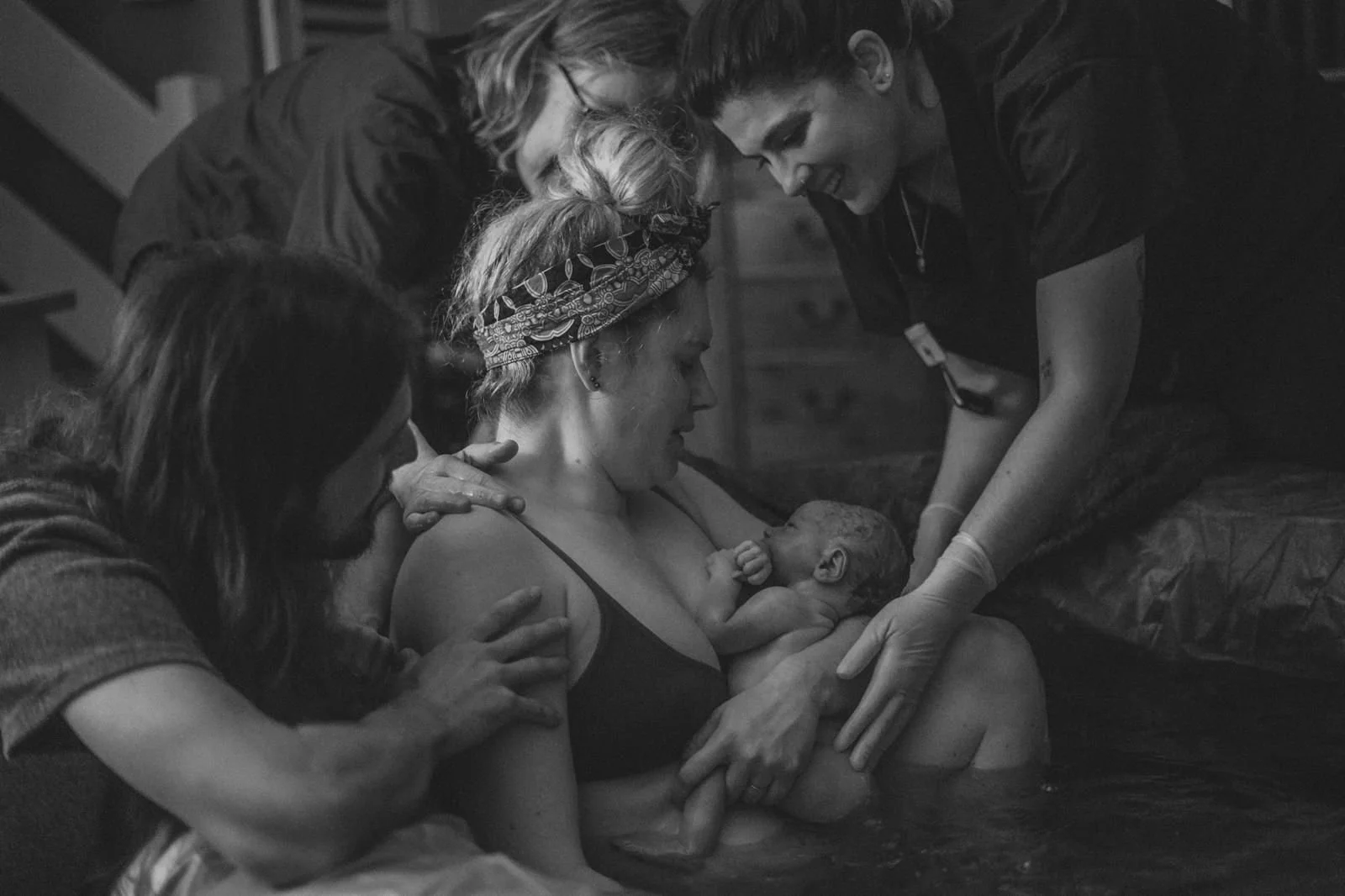 Black and white photo of a woman giving birth in a birthing pool surrounded by three caregivers, all focused on her, with attentive and gentle expressions.