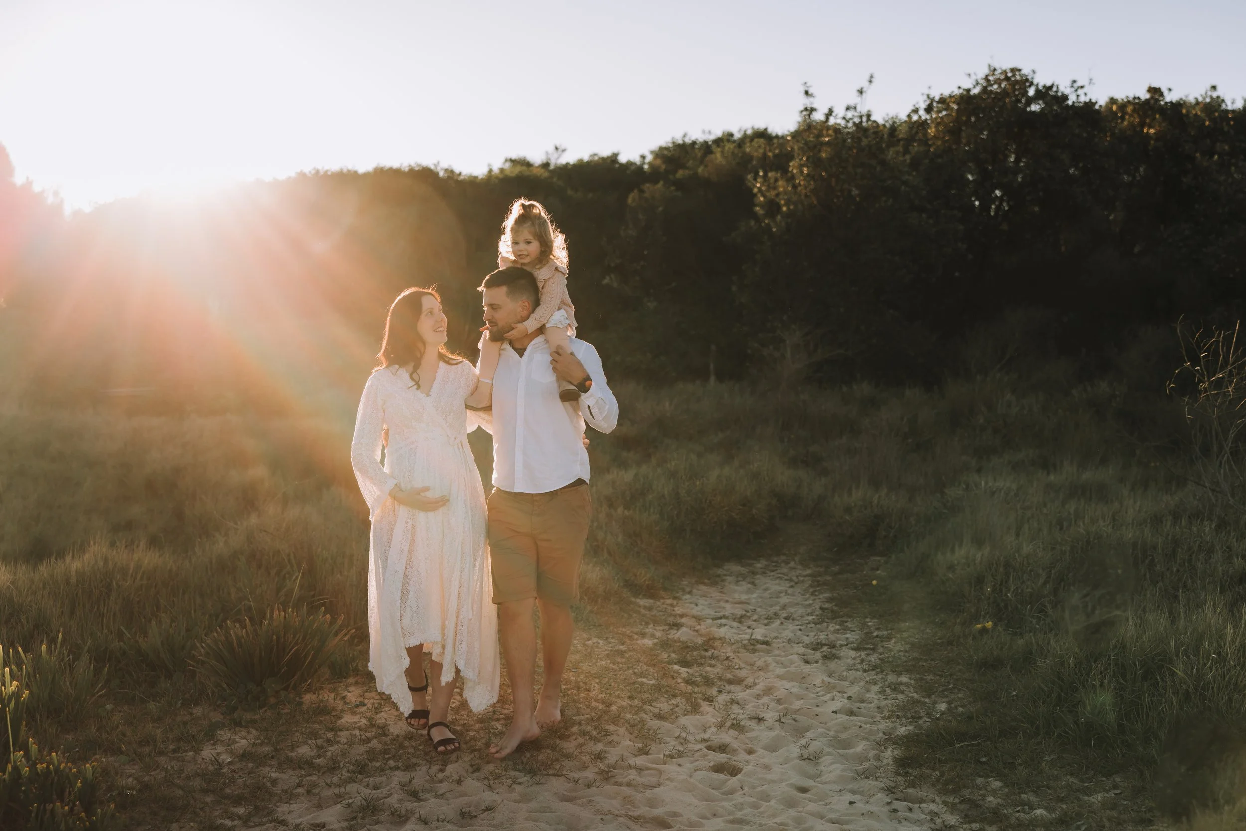 "Sun-drenched family maternity photo of a pregnant woman walking hand-in-hand with her partner, who carries their young daughter on his shoulders, along a grassy path at golden hour — by Nicole Butler Photography at Redhead."