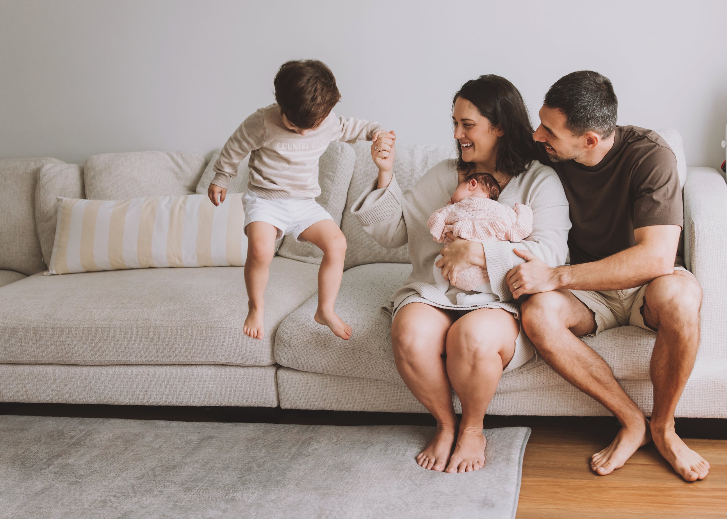A family on a light-colored sofa, with the mother cradling a newborn and the father holding a toddler’s hand as they jump. Photographed by Nicole Butler Photography.