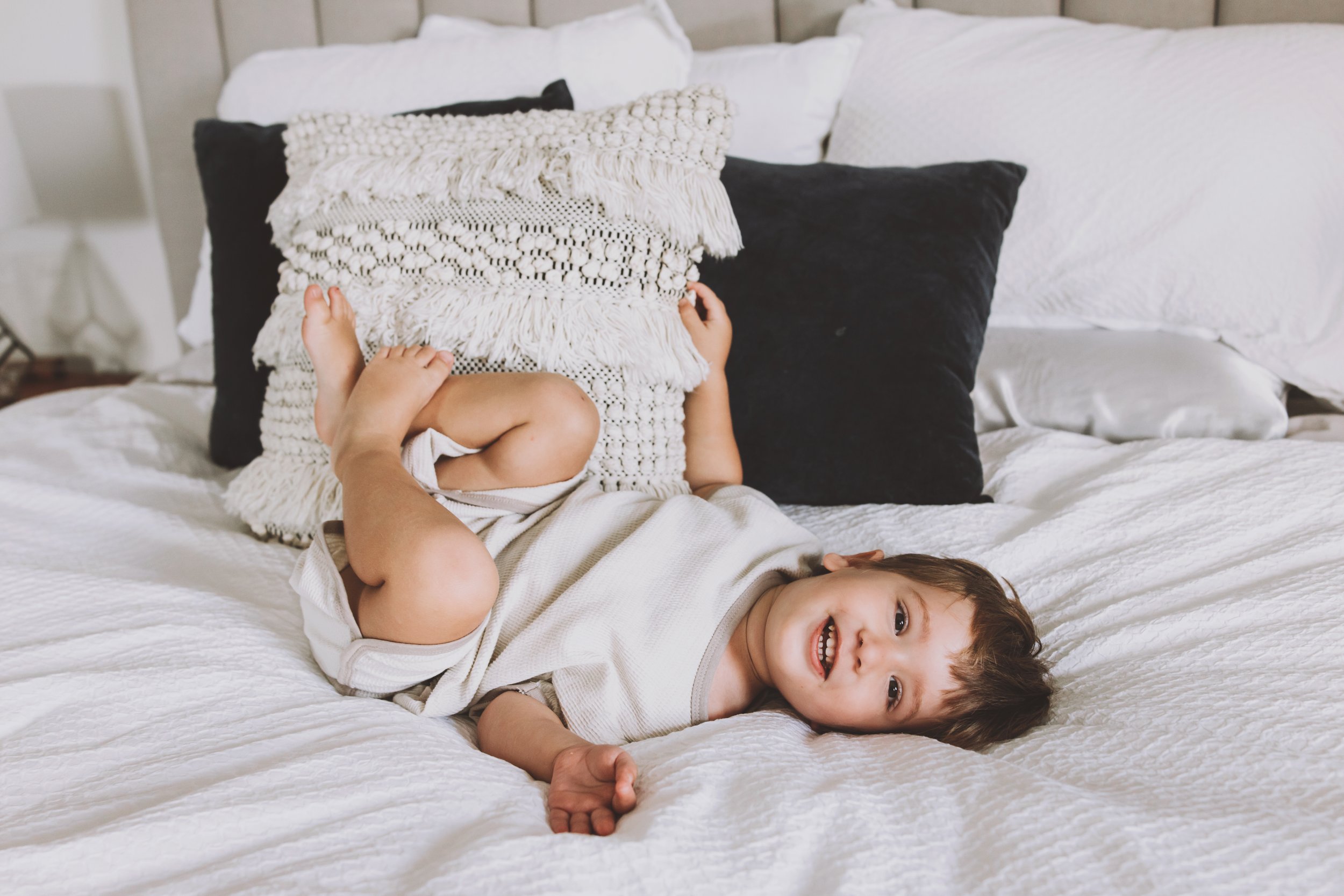 A toddler lying on a white bed, playfully leaning against pillows and smiling. Photographed by Nicole Butler Photography.