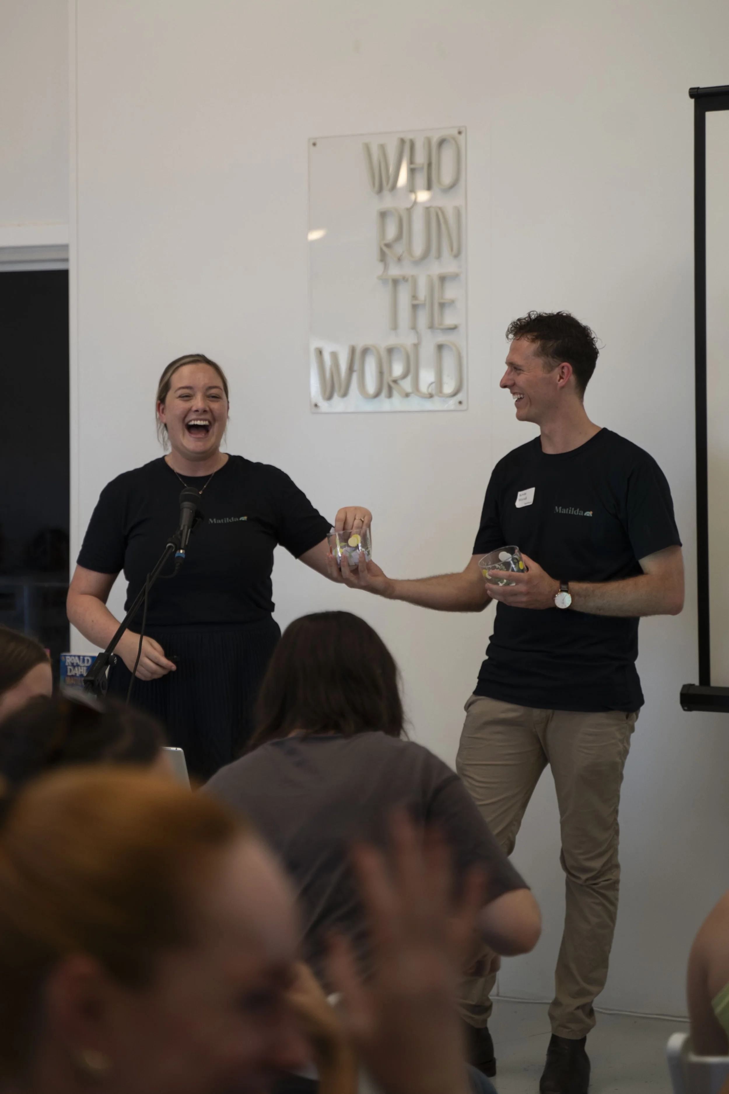 A man and woman laughing and smiling at an event, with the woman holding a clear container with small items, and the man offering her another container. The woman is wearing a black T-shirt, and the man is wearing a black T-shirt and khaki pants. In the background, there is a sign on the wall that says "WHO RUN THE WORLD." There are several people seated in the foreground.