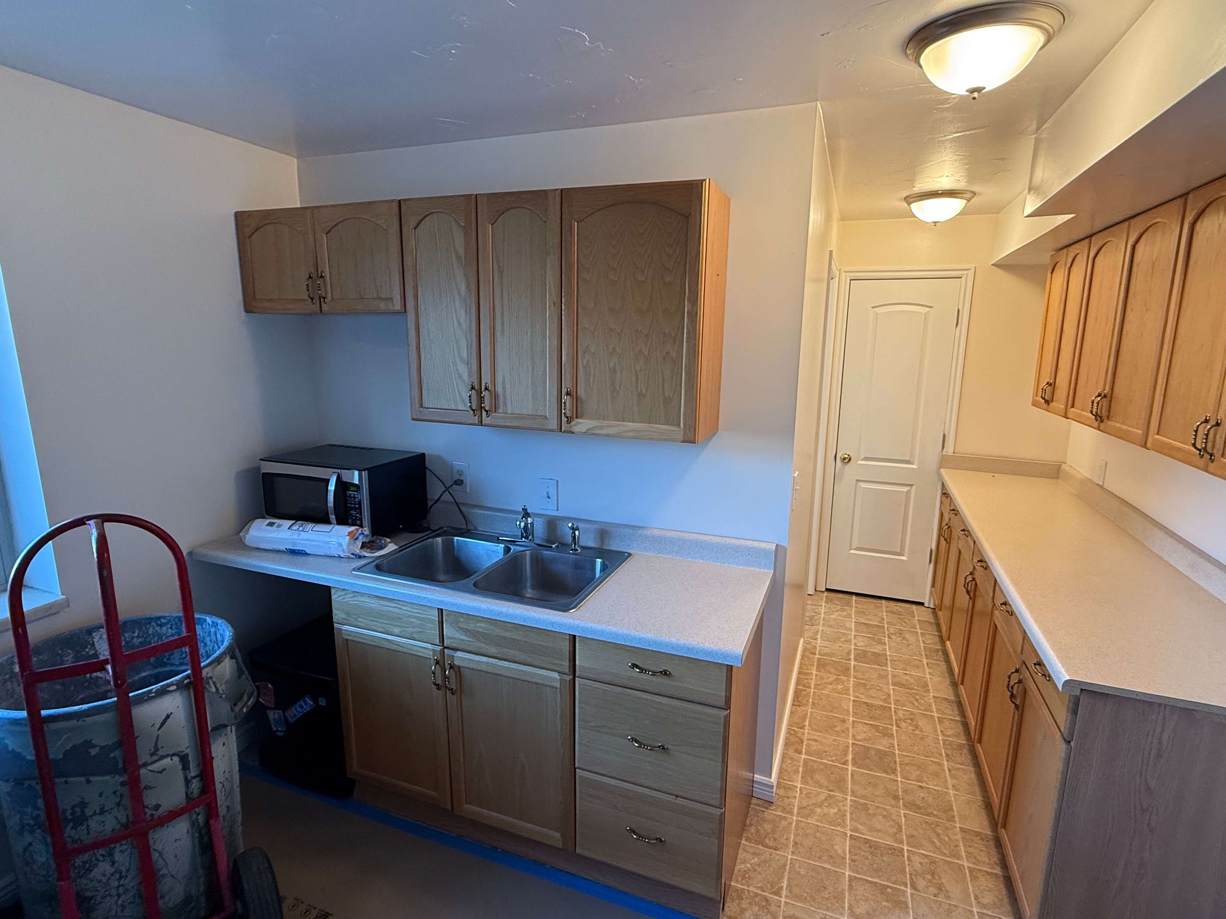 Kitchen with wooden cabinets, a countertop with a sink, a small microwave, and a white door. There are two ceiling lights and a tiled floor. Some objects including a bucket and boxed items are on the countertop.