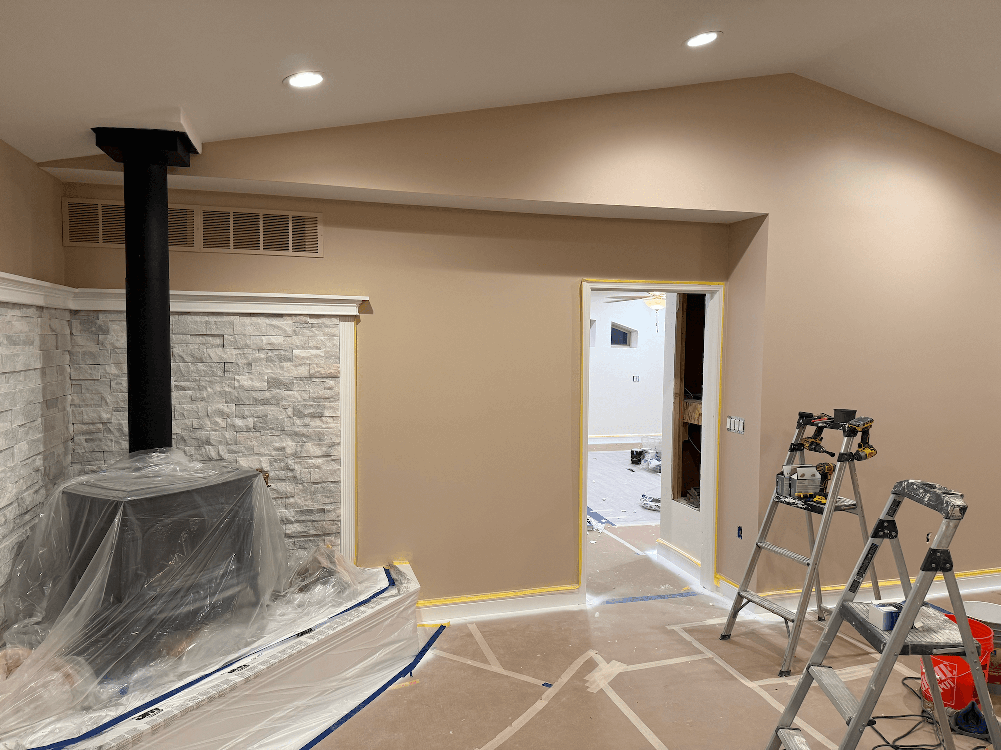 Living room under renovation with plastic sheeting on the floor and walls, a stepped ladder, and construction tools. A black chimney pipe extends from the ceiling towards a stone accent wall. The doorway frames are taped off, revealing an adjoining room.