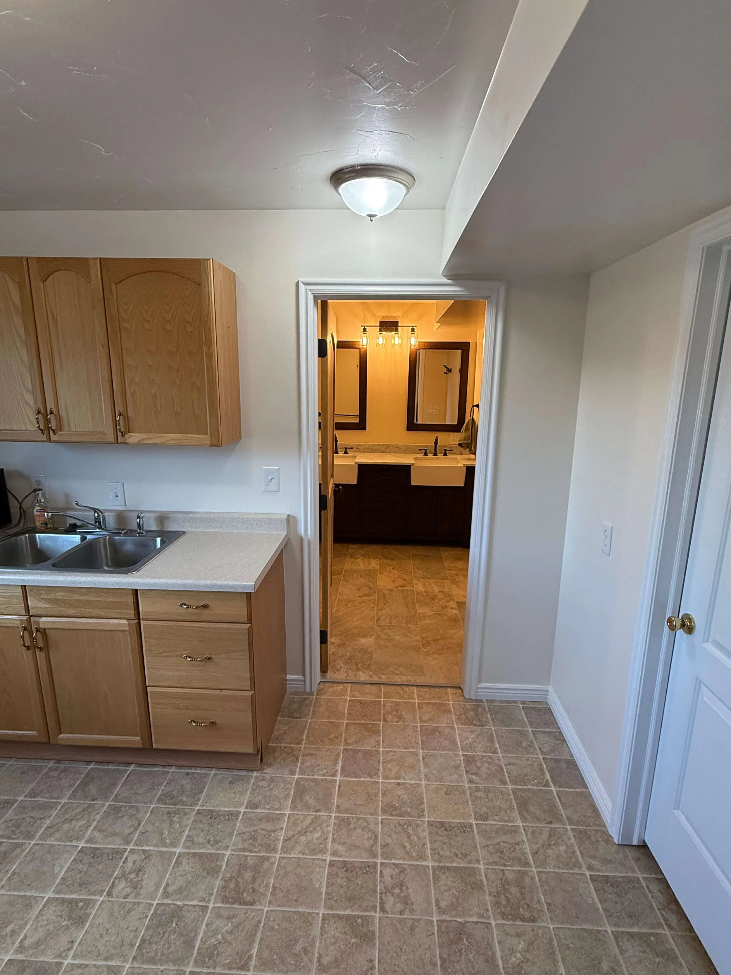 Interior view of a kitchen leading into a bathroom area with two sinks, a mirror, and a lighting fixture, with tiled floor and wooden cabinets.