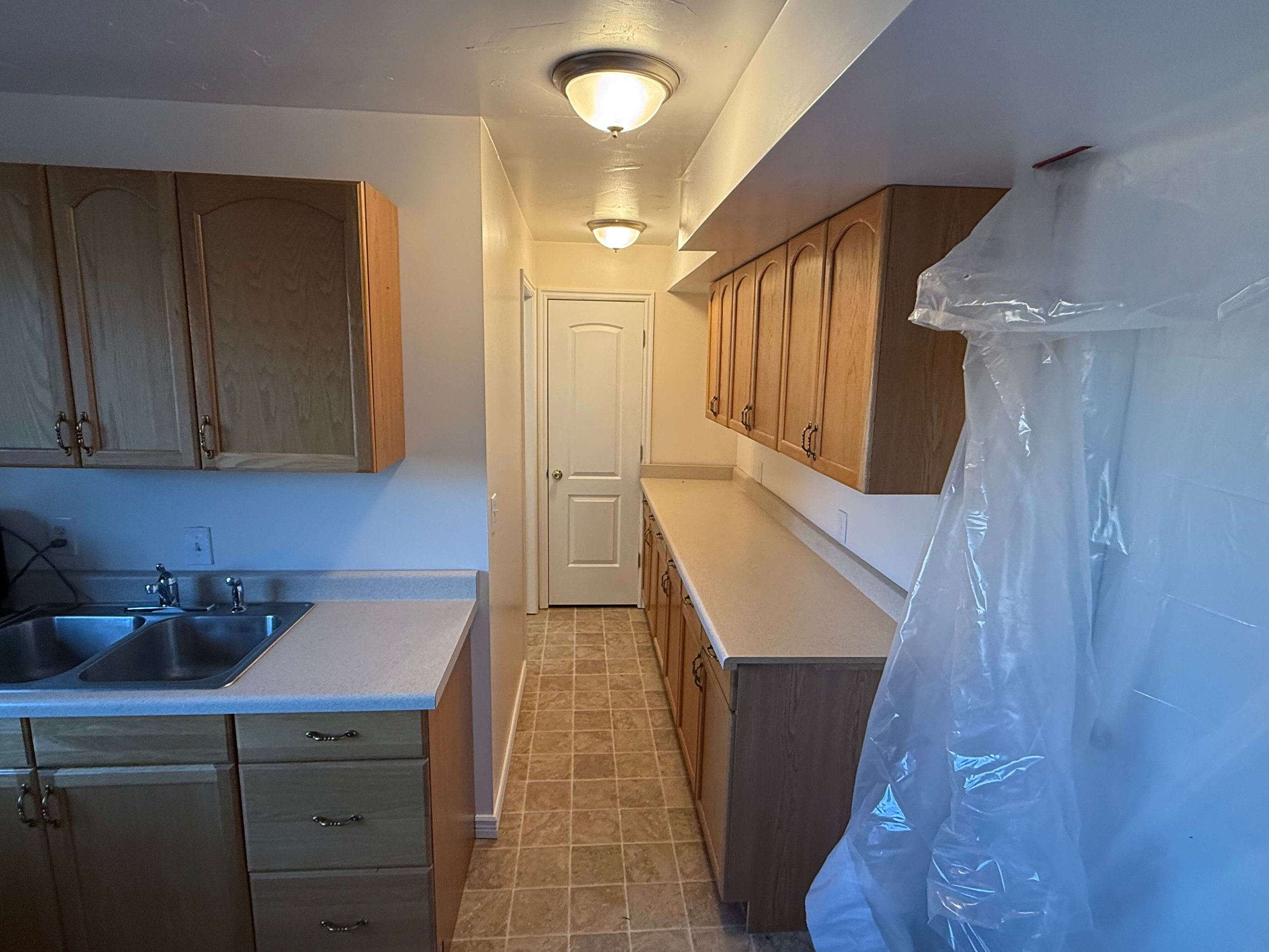 A kitchen with wooden cabinets, beige countertops, a double sink, and a tiled floor. The ceiling has two light fixtures. A door at the end of the kitchen leads to another room, and a clear plastic bag is placed on the counter on the right side.
