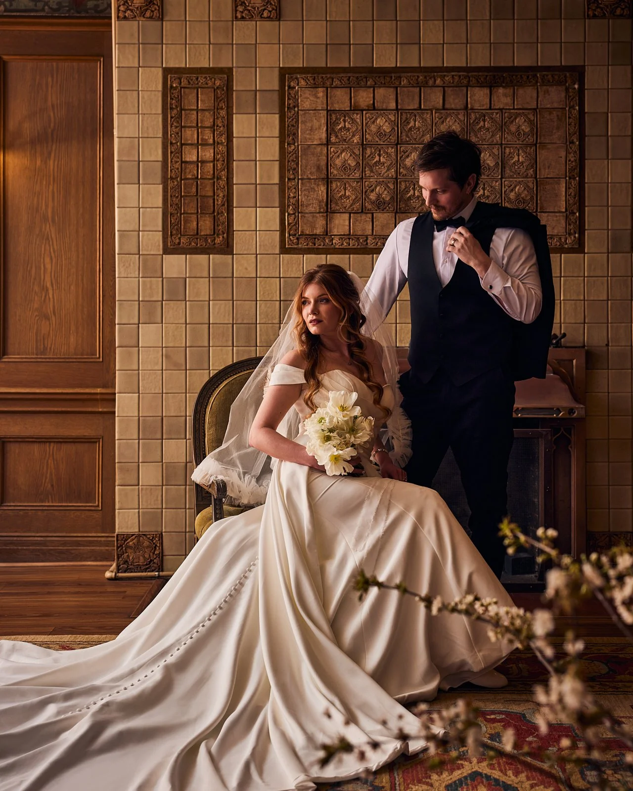 Bride seated in a vintage chair in a wedding gown holding a bouquet of white flowers, with a groom standing beside her adjusting his bow tie, in a warmly lit room with tiled wall and ornate wood paneling.