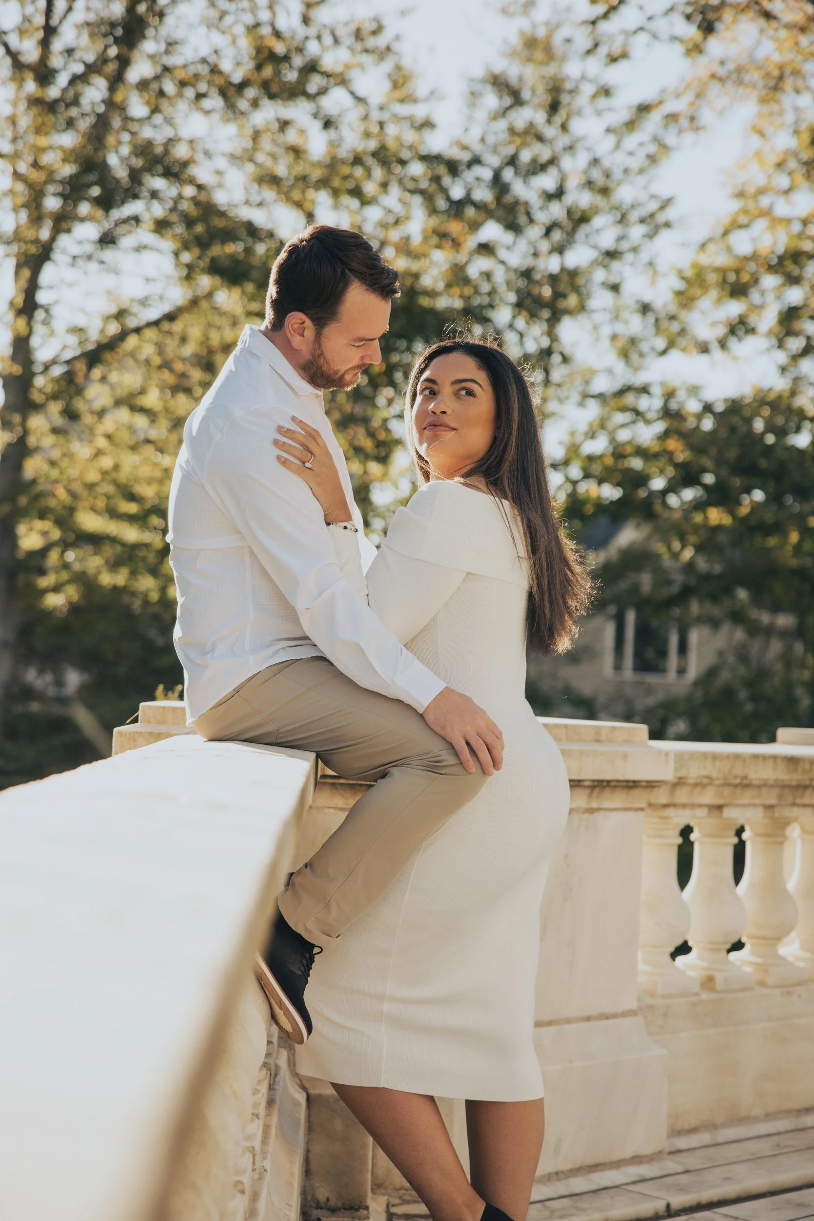 A couple sitting on a balustrade outdoors during daytime, with trees in the background. They are dressed in white and beige and are gazing at each other lovingly.