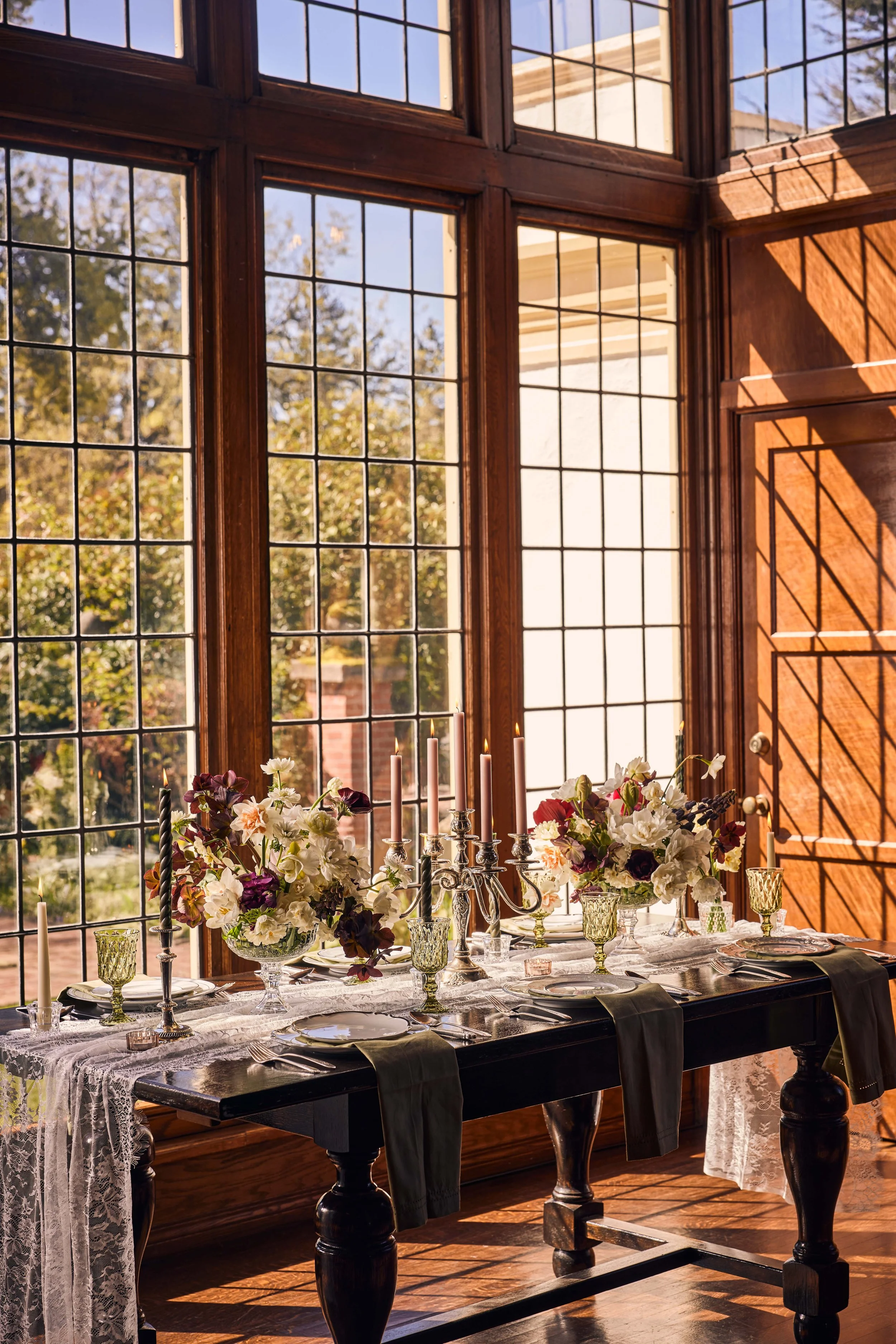 Sunlight streams through large wooden window panes illuminating a decorated table with floral arrangements, candles, and fine dining ware set for a meal in a room with wooden walls and flooring.