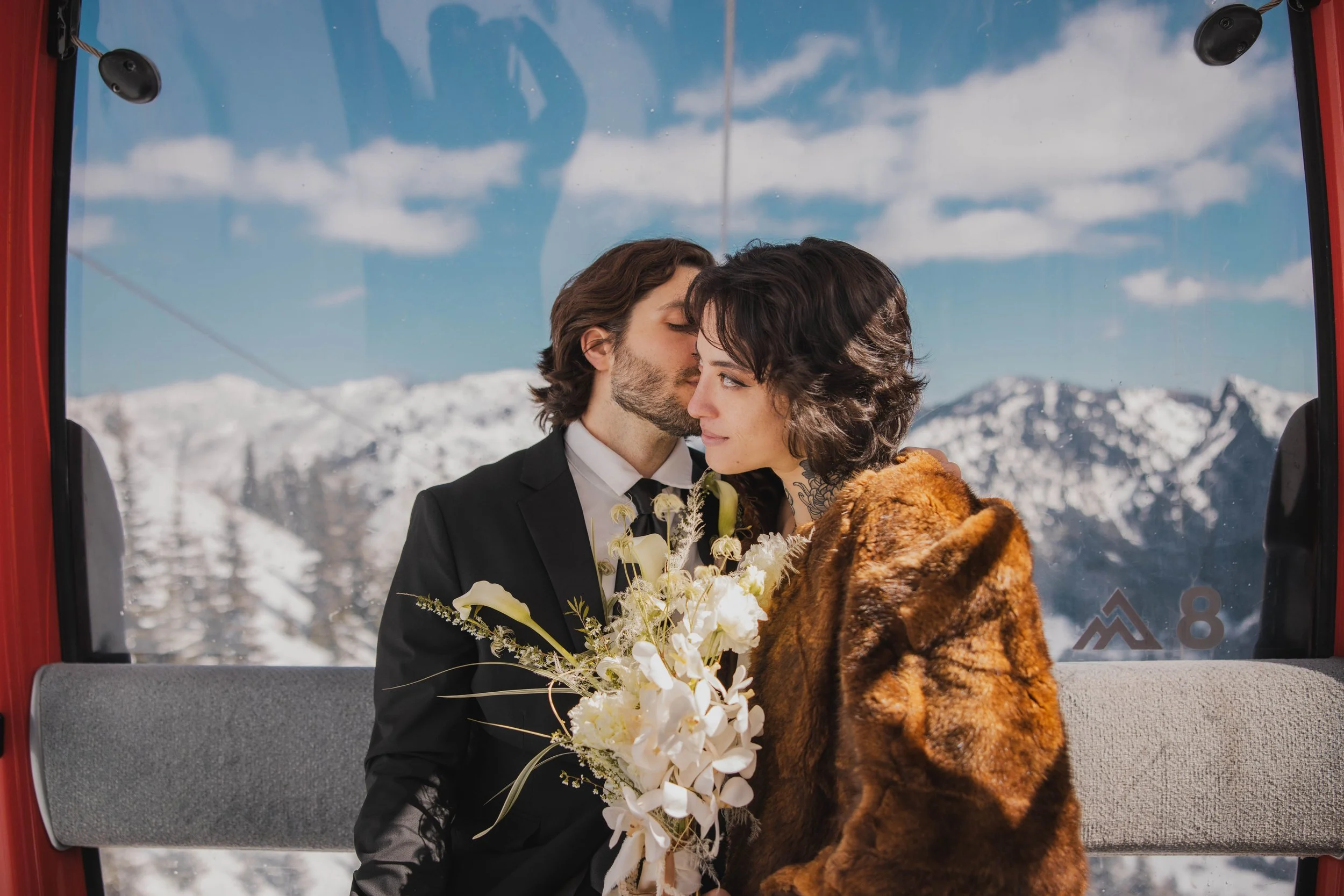 A couple sharing a kiss while riding a ski lift with snowy mountains and blue sky in the background. The man is in formal attire, the woman is wearing a fur coat, and she is holding a bouquet of white flowers.
