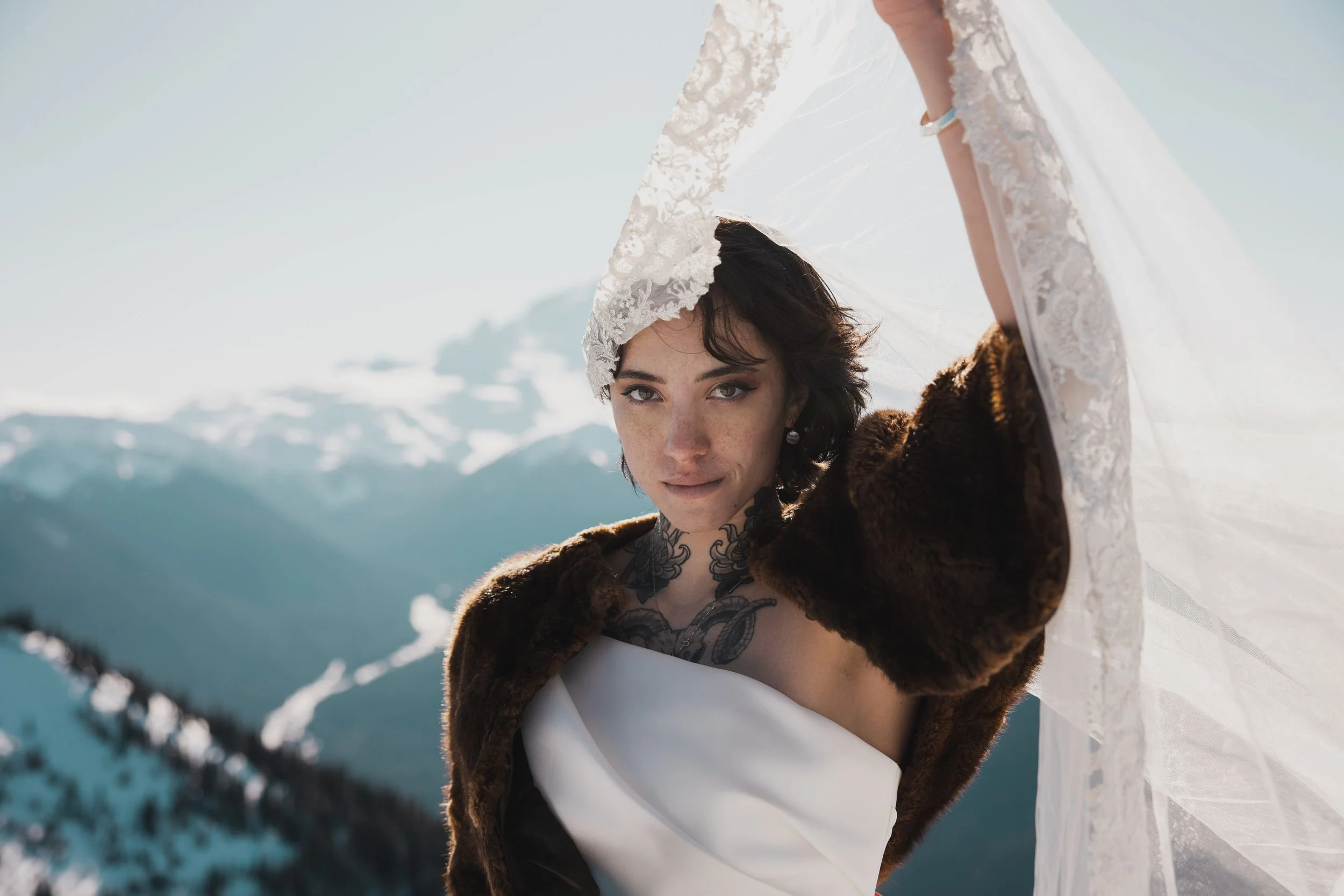 A woman with short dark hair and tattoos on her chest, wearing a white dress and a fur coat, holds a white lace wedding veil above her head in a snowy mountain landscape.