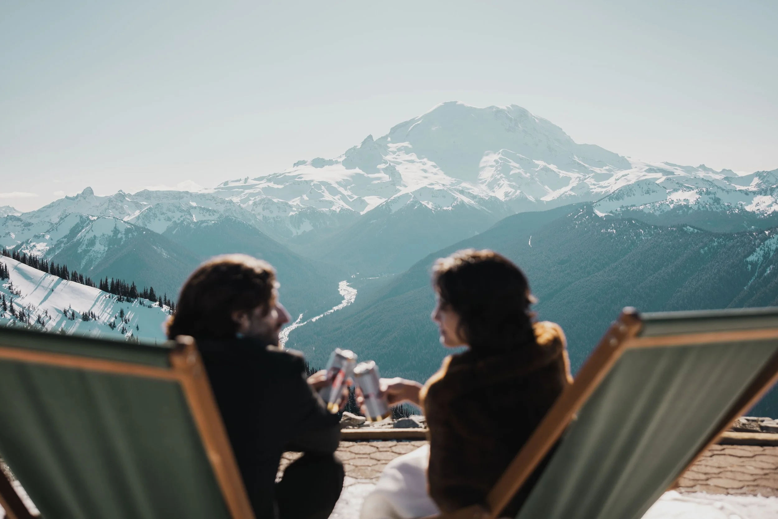 Two people sitting on lounge chairs facing a snow-capped mountain range, with a prominent mountain in the center, under a clear sky.