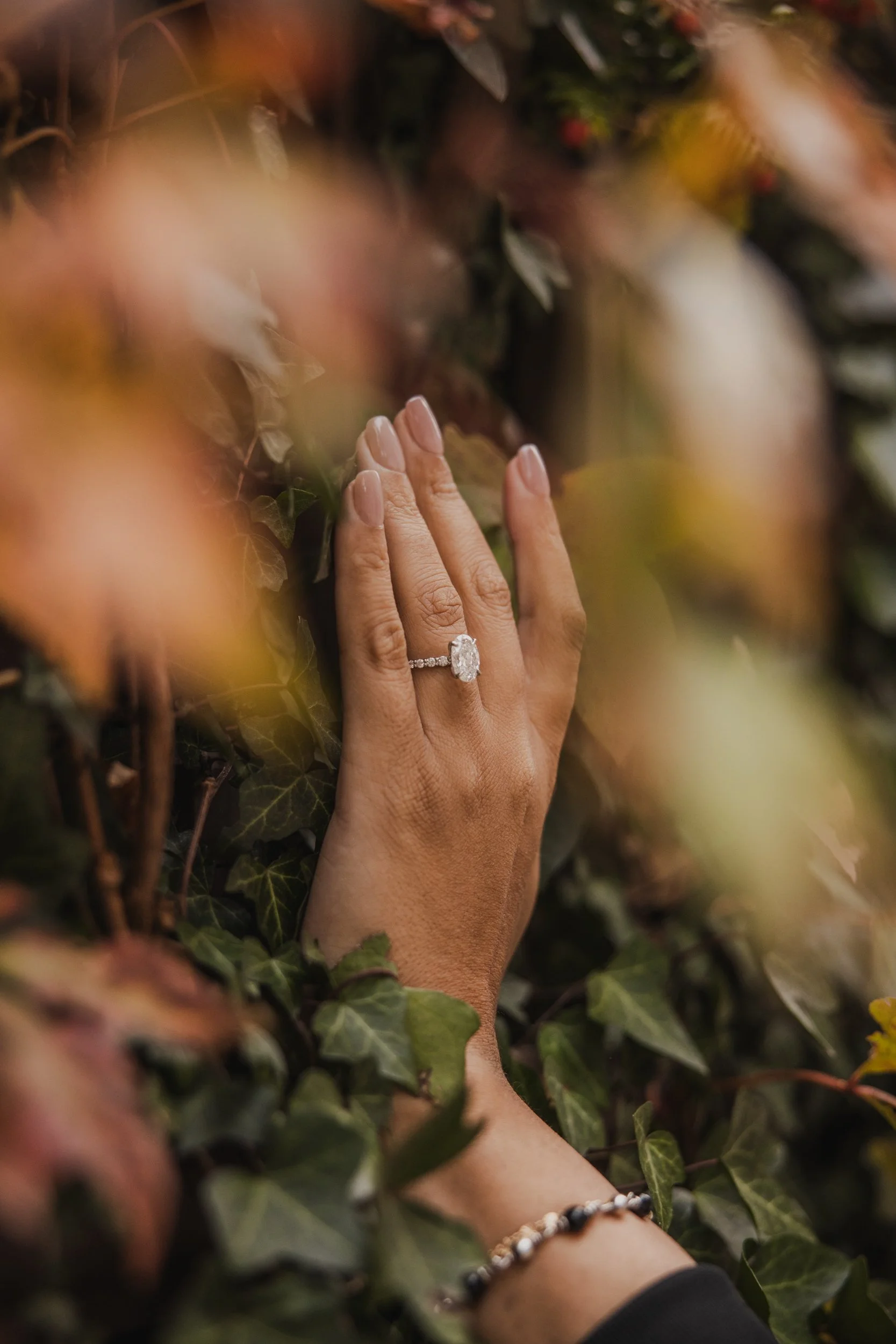 A woman’s hand with an engagement ring resting on green and brown leaves.