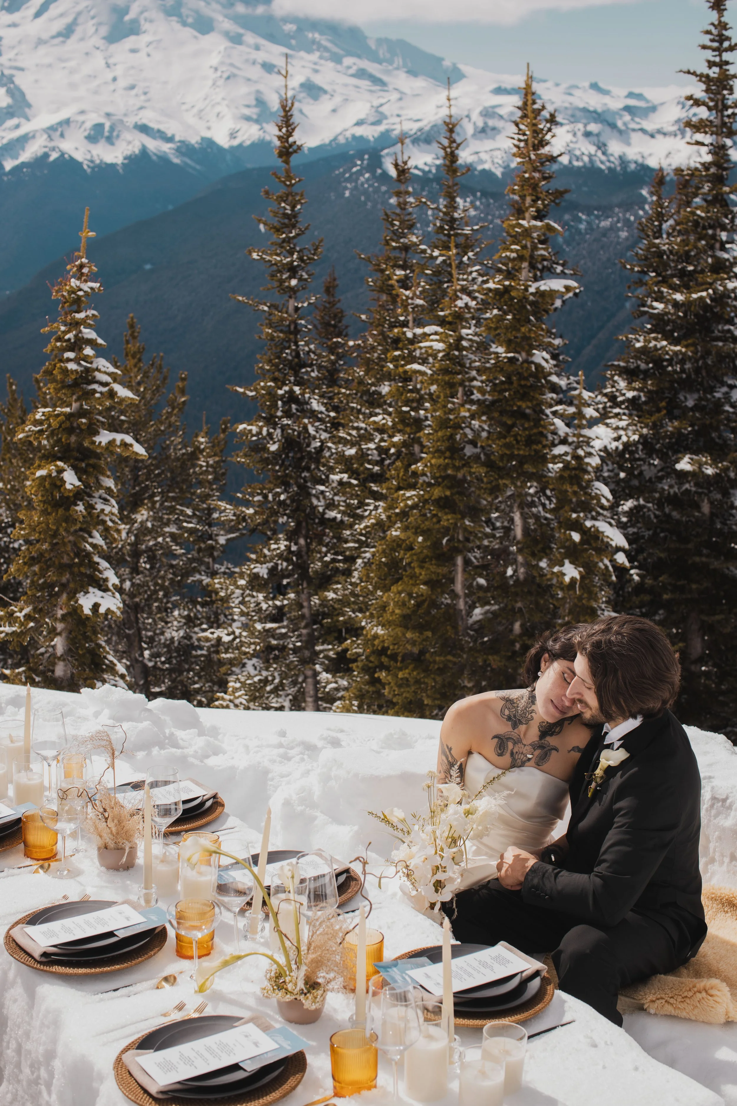 A couple having a romantic moment at a wedding reception outdoors in a snowy mountain setting. The bride and groom are sitting close together, surrounded by a beautifully decorated table with candles, flowers, and elegant tableware.
