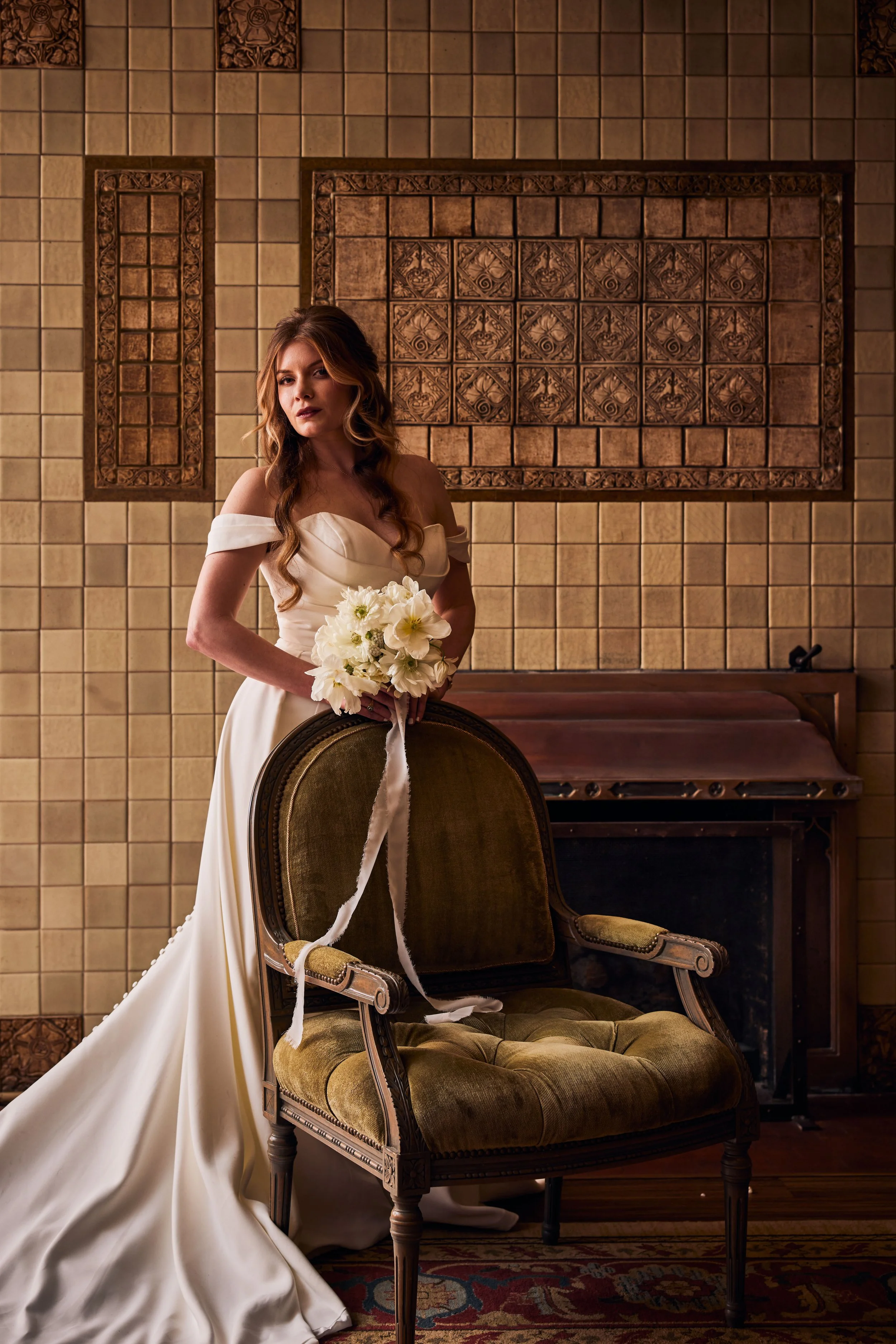 A woman in a white off-shoulder wedding dress holding a bouquet of white flowers, standing next to a vintage green velvet armchair in front of a decorative tiled wall.