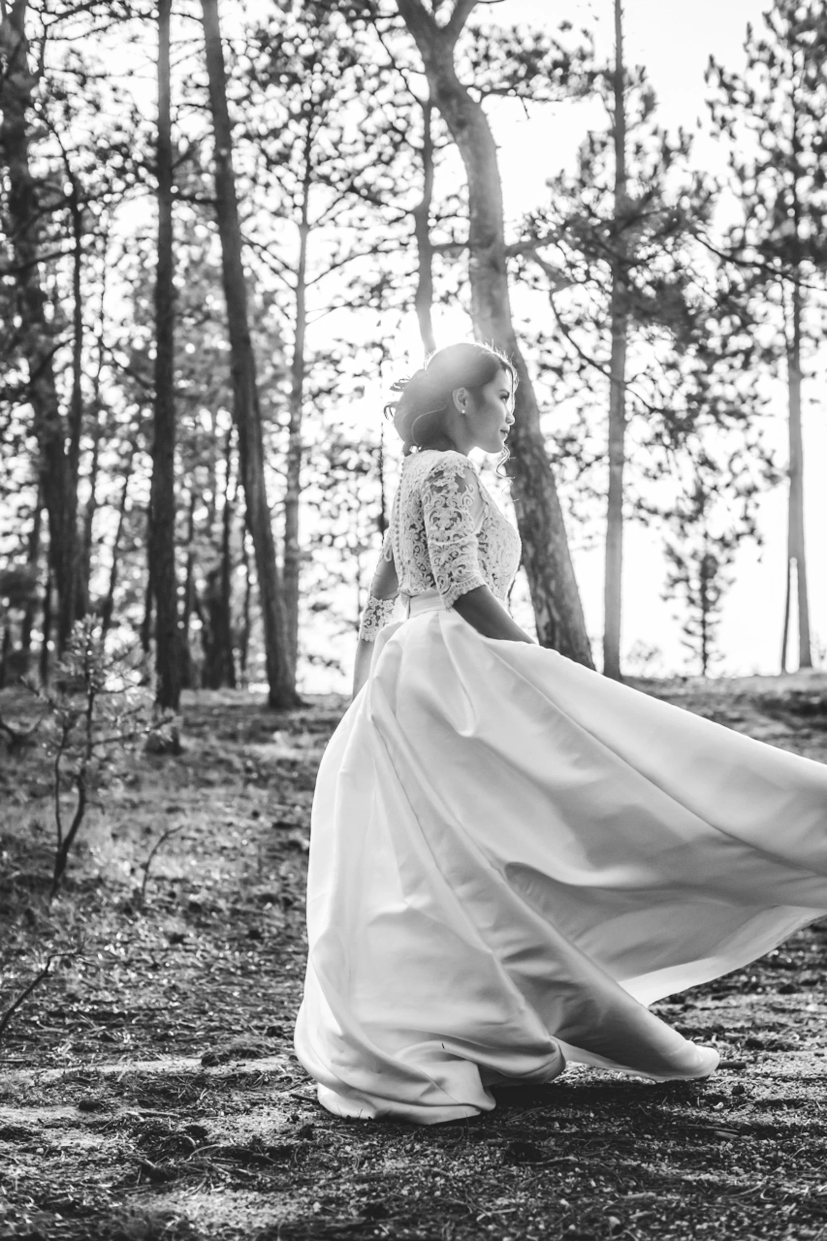 Black and white photo of a woman in a wedding dress walking in a forest, with trees in the background and sunlight filtering through.