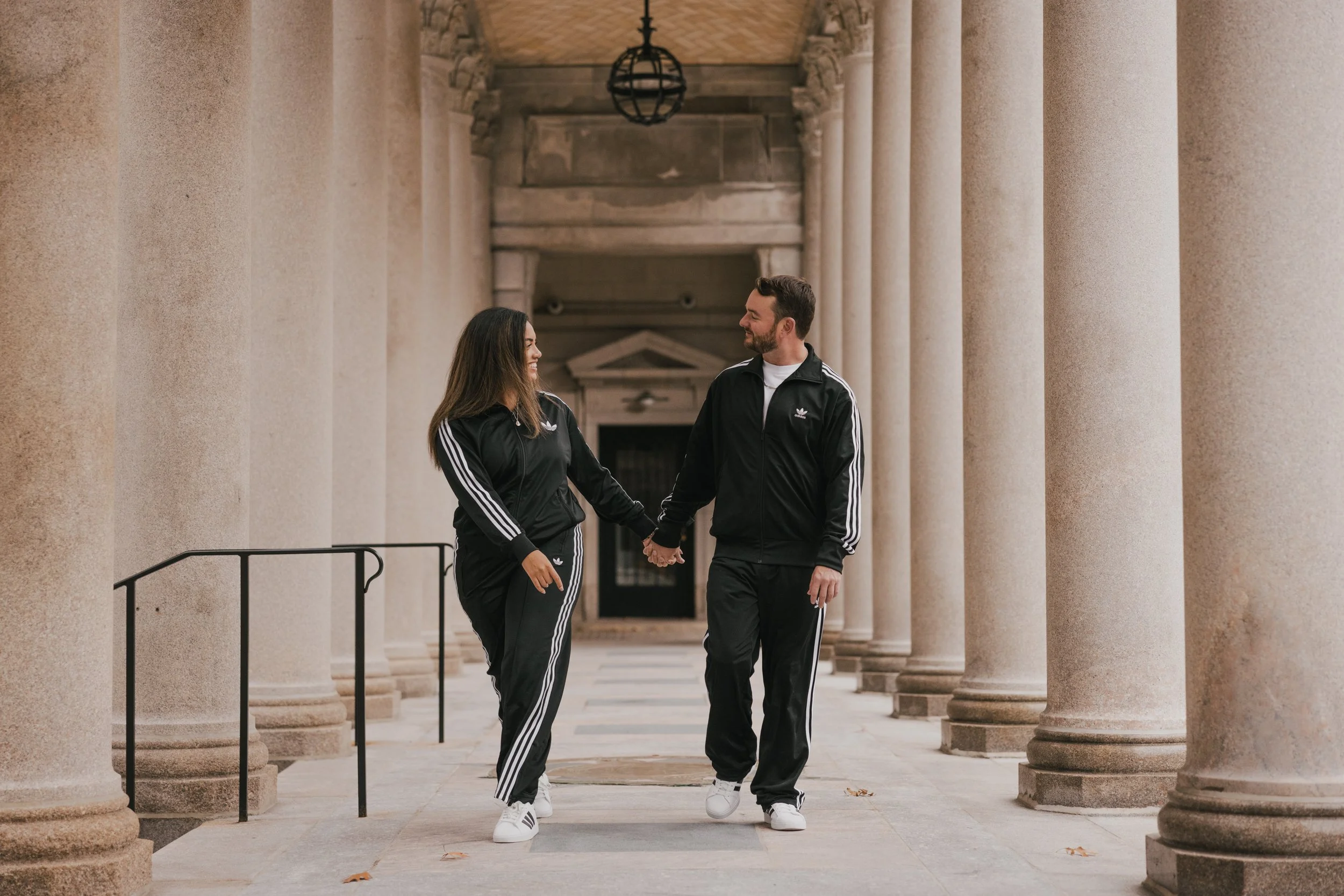 A man and woman dressed in black Adidas tracksuits holding hands and smiling at each other while walking through a colonnade with tall stone columns.