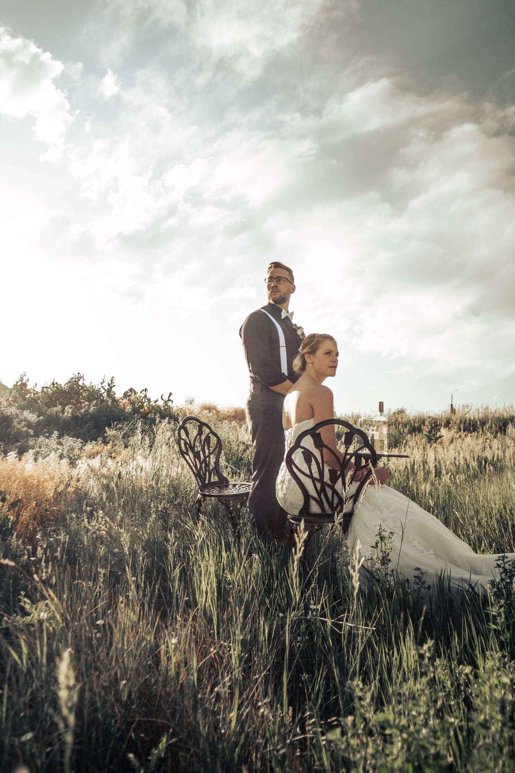 A wedding couple is outdoors in a grassy field during sunset. The bride is sitting at a small table with a bottle of alcohol and glasses, wearing a strapless wedding gown. The groom stands behind her, wearing a dark suit with suspenders and a bow tie