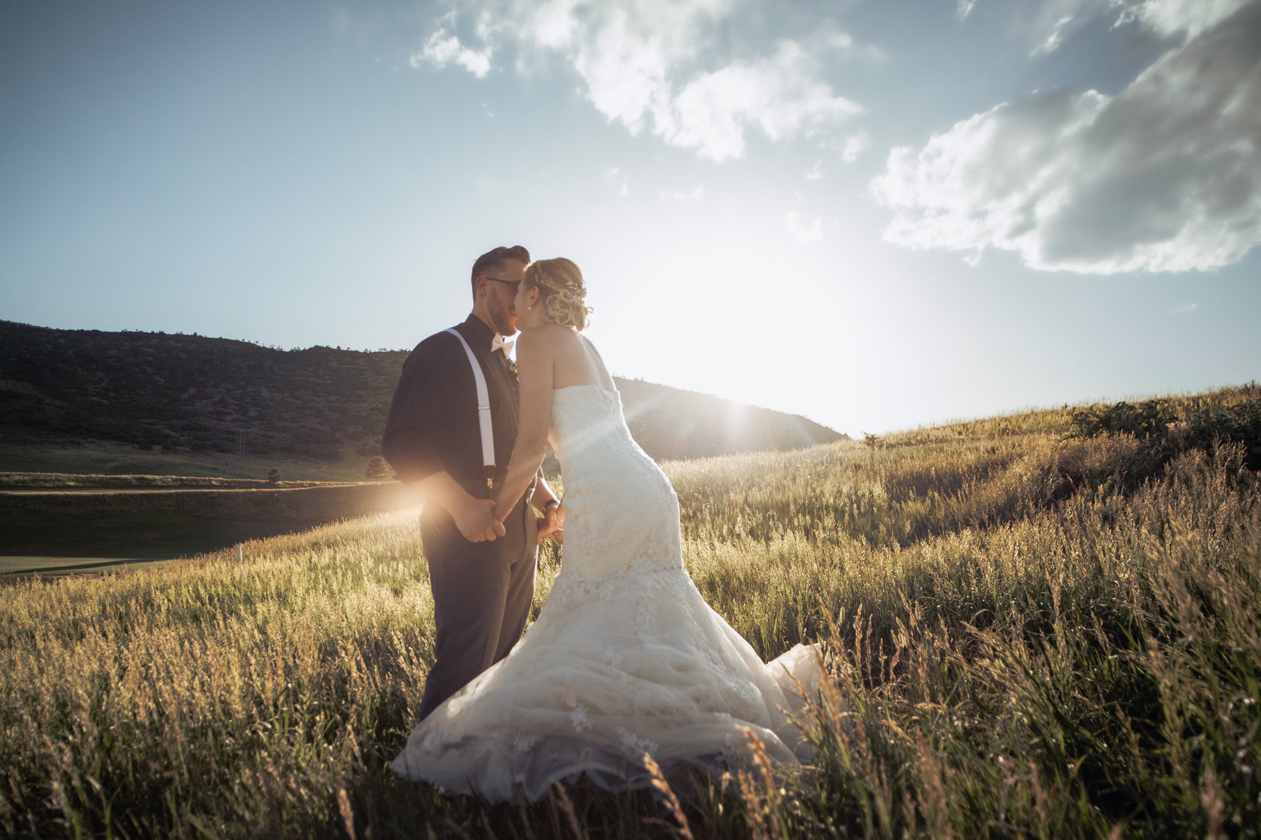 A newlywed couple, a man in a black suit with suspenders and a woman in a white wedding dress, sharing a kiss in a field during sunset with hills in the background.