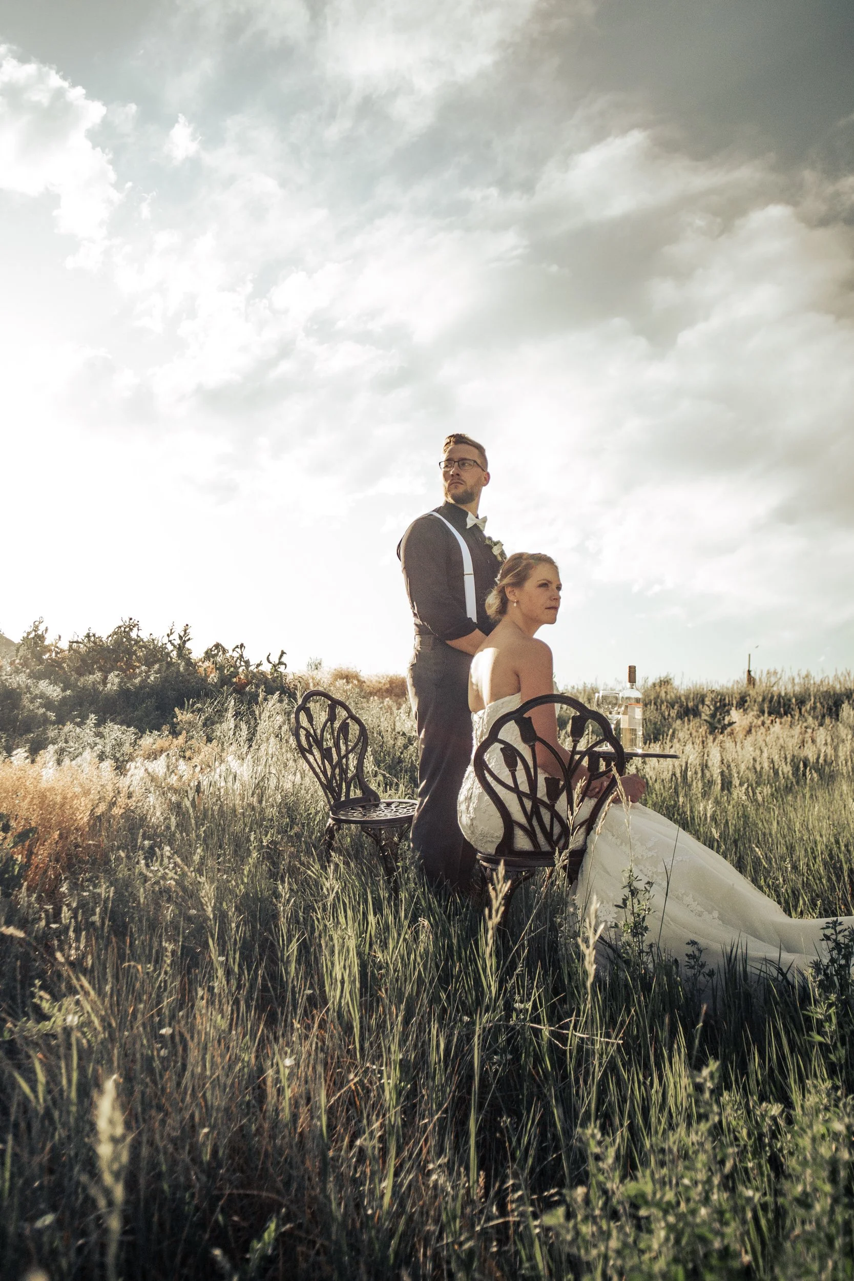 A couple dressed in wedding attire outdoors in a grassy field under a partly cloudy sky. One person is standing, wearing glasses and a black tuxedo with suspenders, while the other is seated in a white wedding dress holding a drink. There is a small 