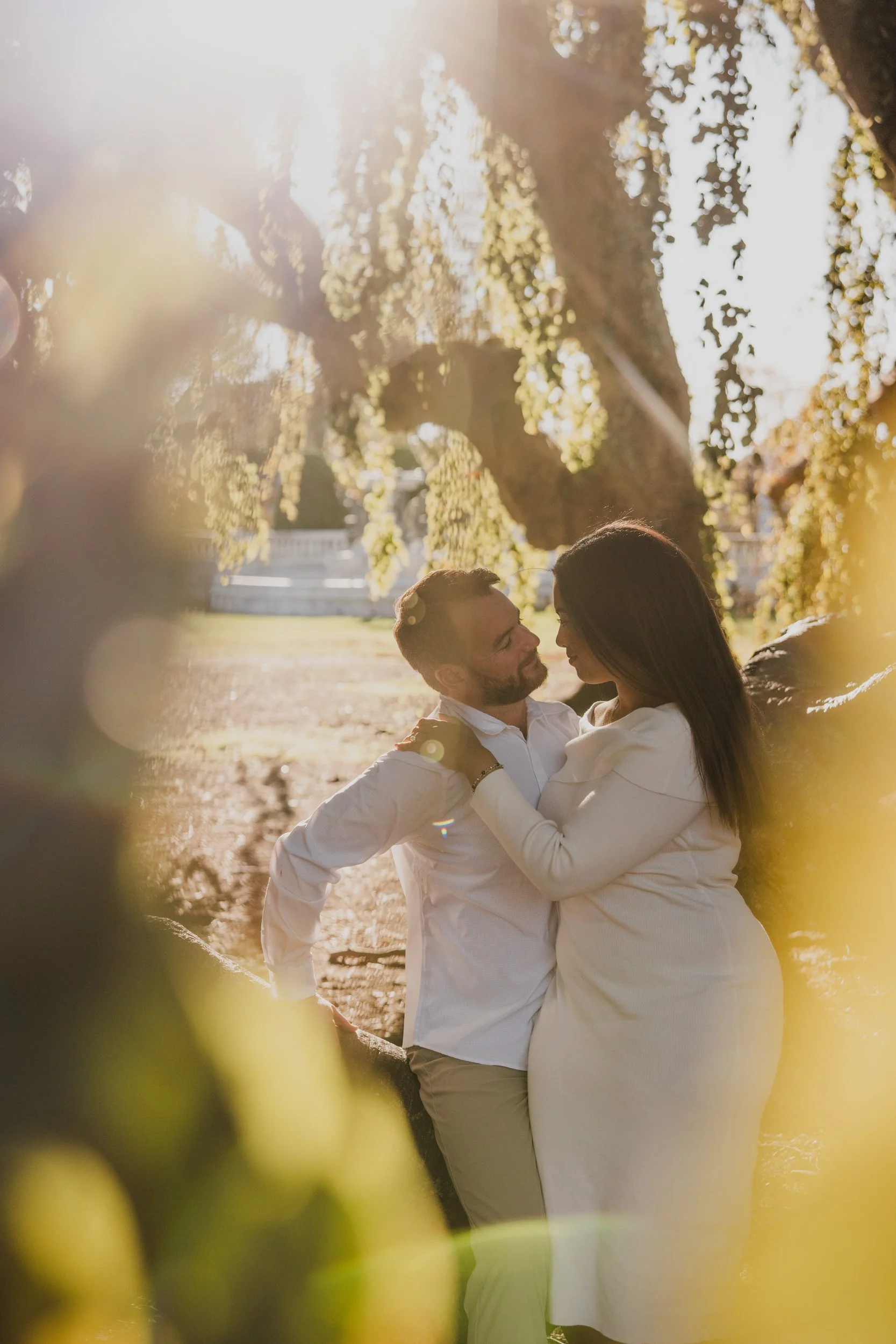 A couple dressed in white, standing close and facing each other beneath a large tree in a park at sunset, with warm sunlight and blurred greenery in the background.