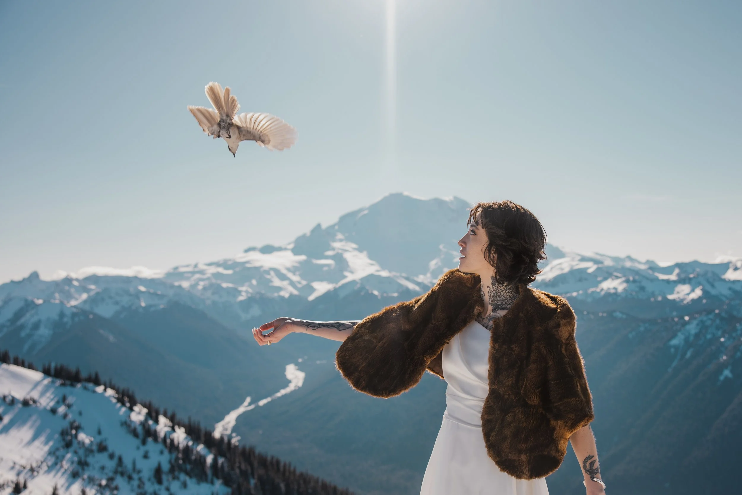 A woman with tattooed arms and short hair standing outdoors in snowy mountains, wearing a white dress and a fur coat, with a bird flying above her toward the sky.
