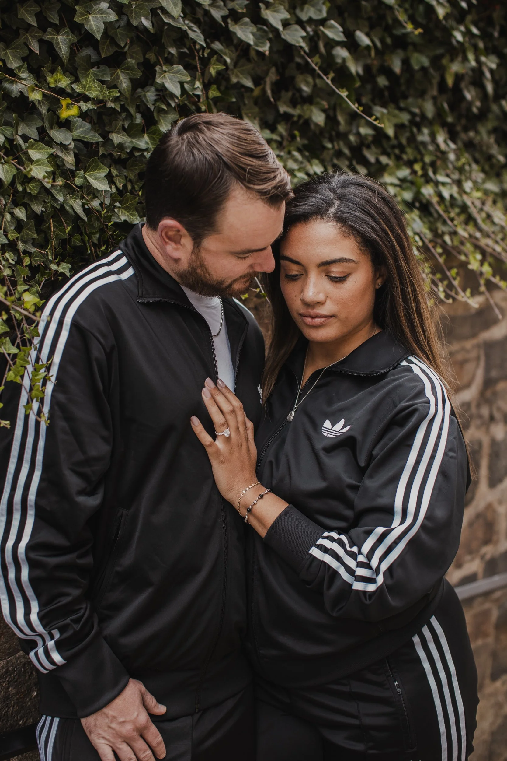 A couple standing close together against a leafy background, wearing matching black Adidas jackets with white stripes, with the woman showing her engagement ring.
