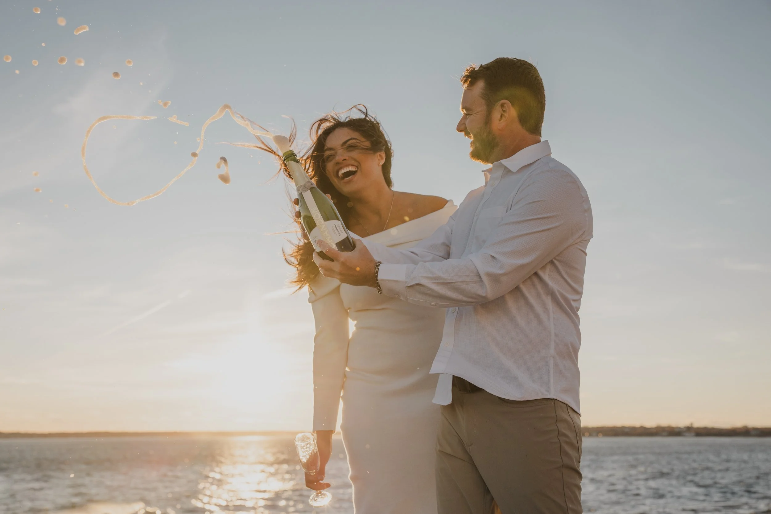 A man and woman celebrating by the water at sunset, opening a bottle of champagne with champagne foam and bubbles flying