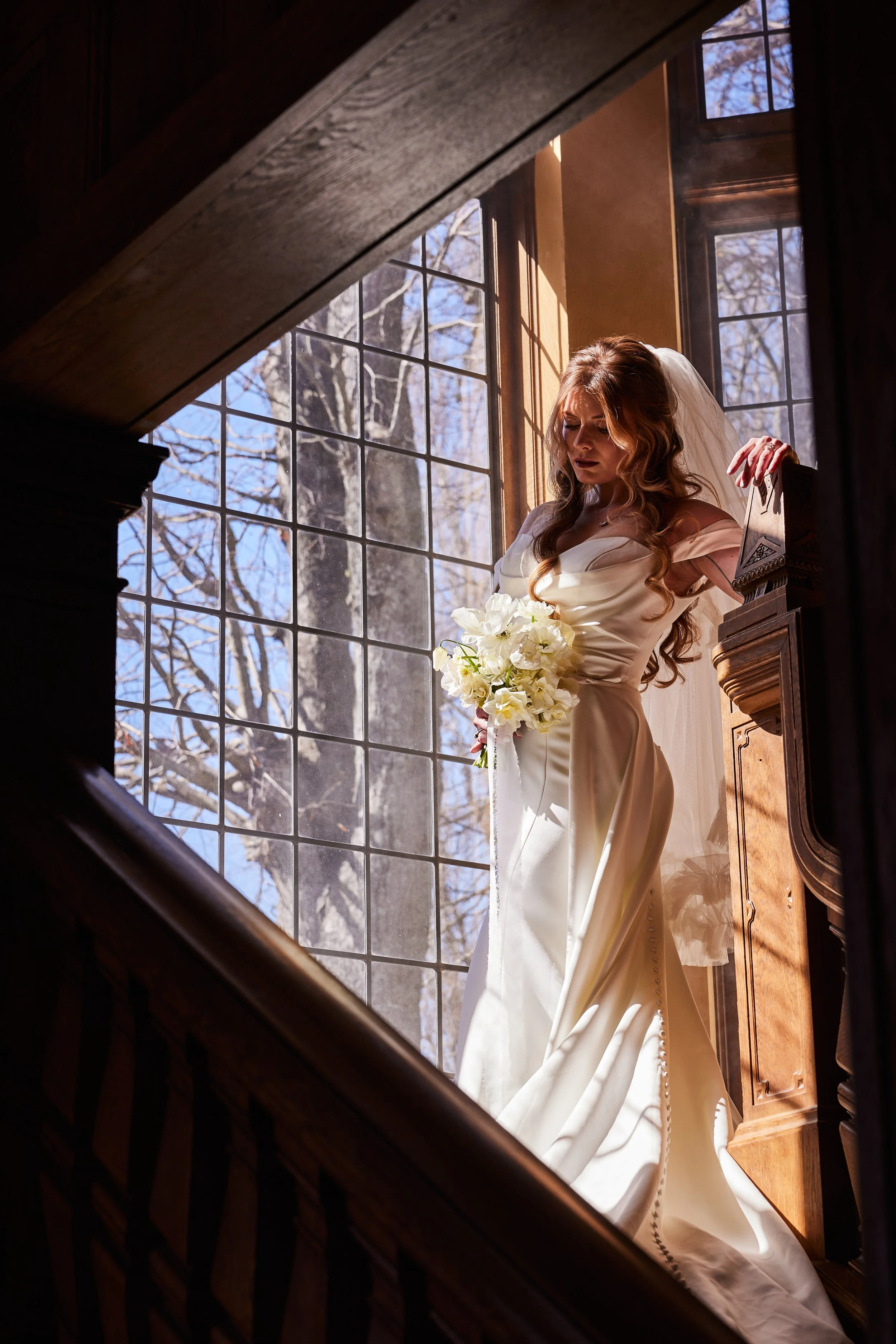A bride in a wedding gown standing indoors near a large window, holding a bouquet of white flowers, with sunlight streaming through the window.