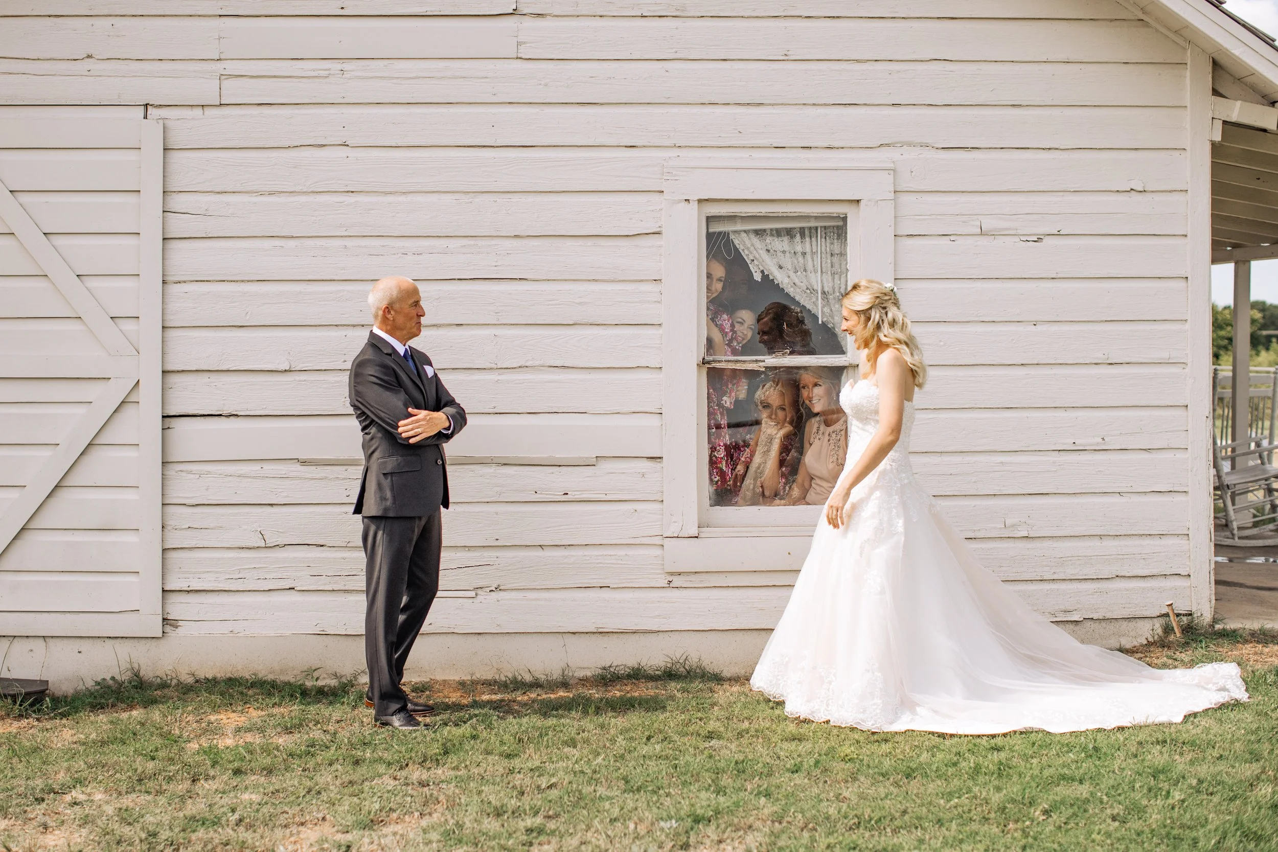 A bride in a white wedding gown standing outside a white wooden barn, looking at an older man in a black suit, while five women look out of a window and observe the scene.