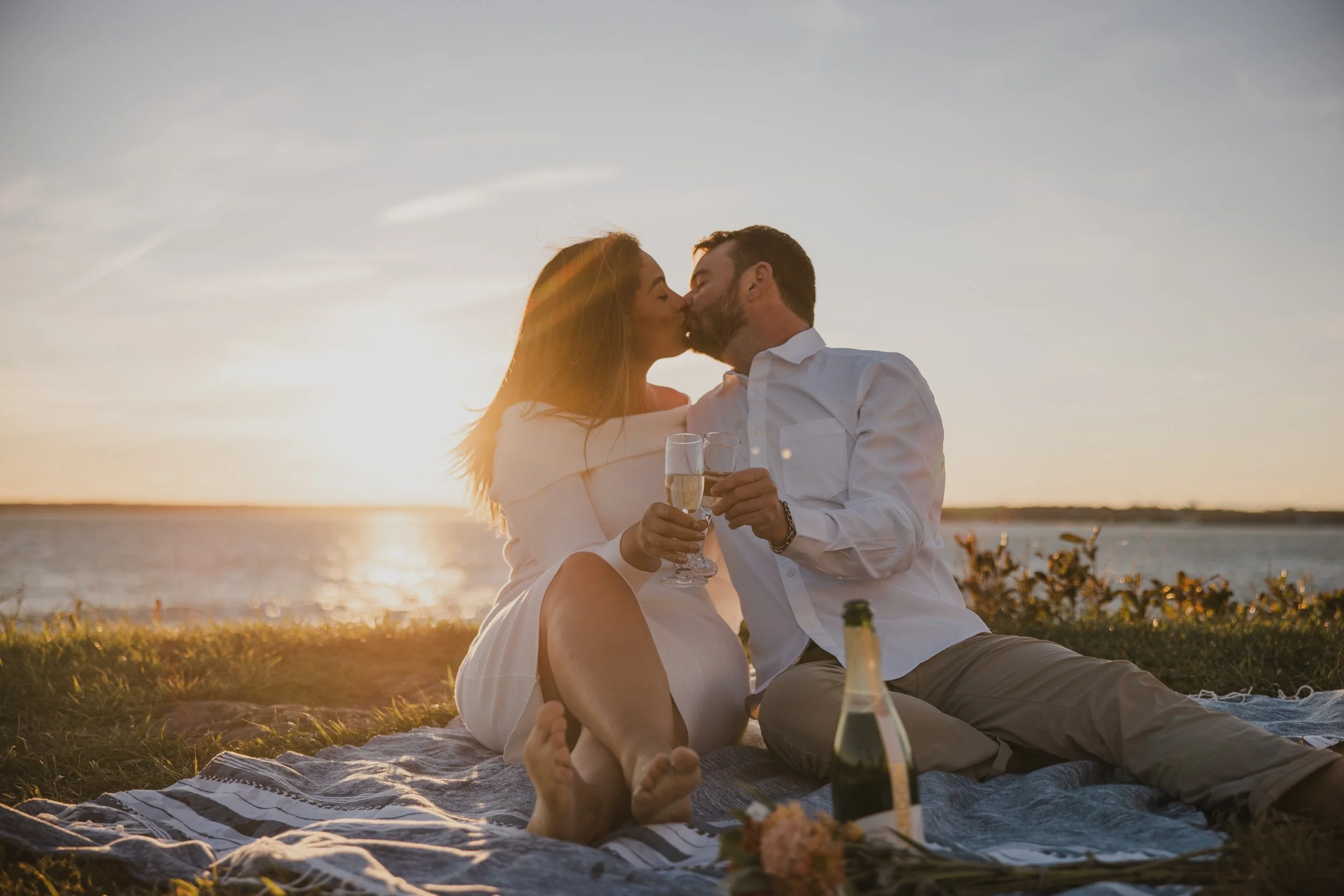 A couple kissing during sunset by the water, sitting on a picnic blanket with champagne glasses and a bottle.