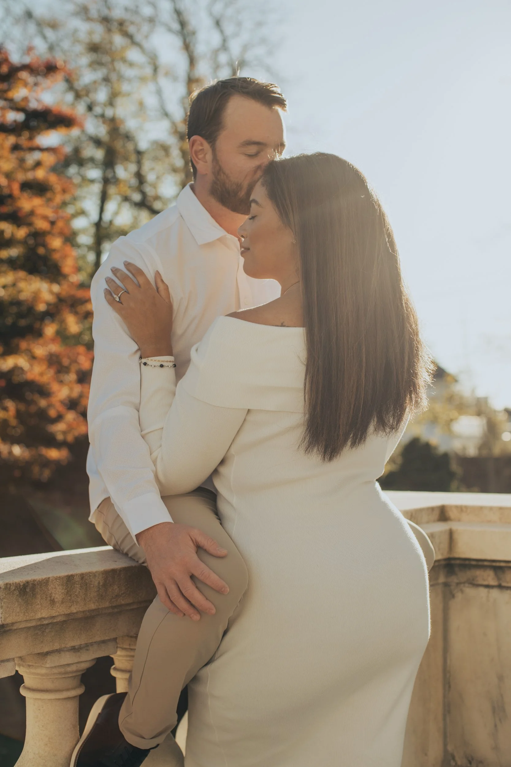 A couple dressed in white clothing embracing outdoors with sunlight in the background, standing near a stone balustrade.