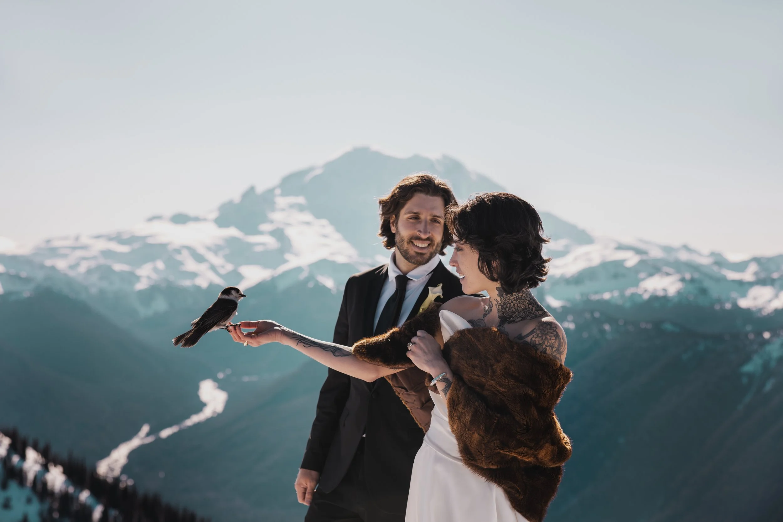 A couple dressed in wedding attire standing outdoors in snowy mountain landscape, with the woman holding a bird on her hand and both smiling.