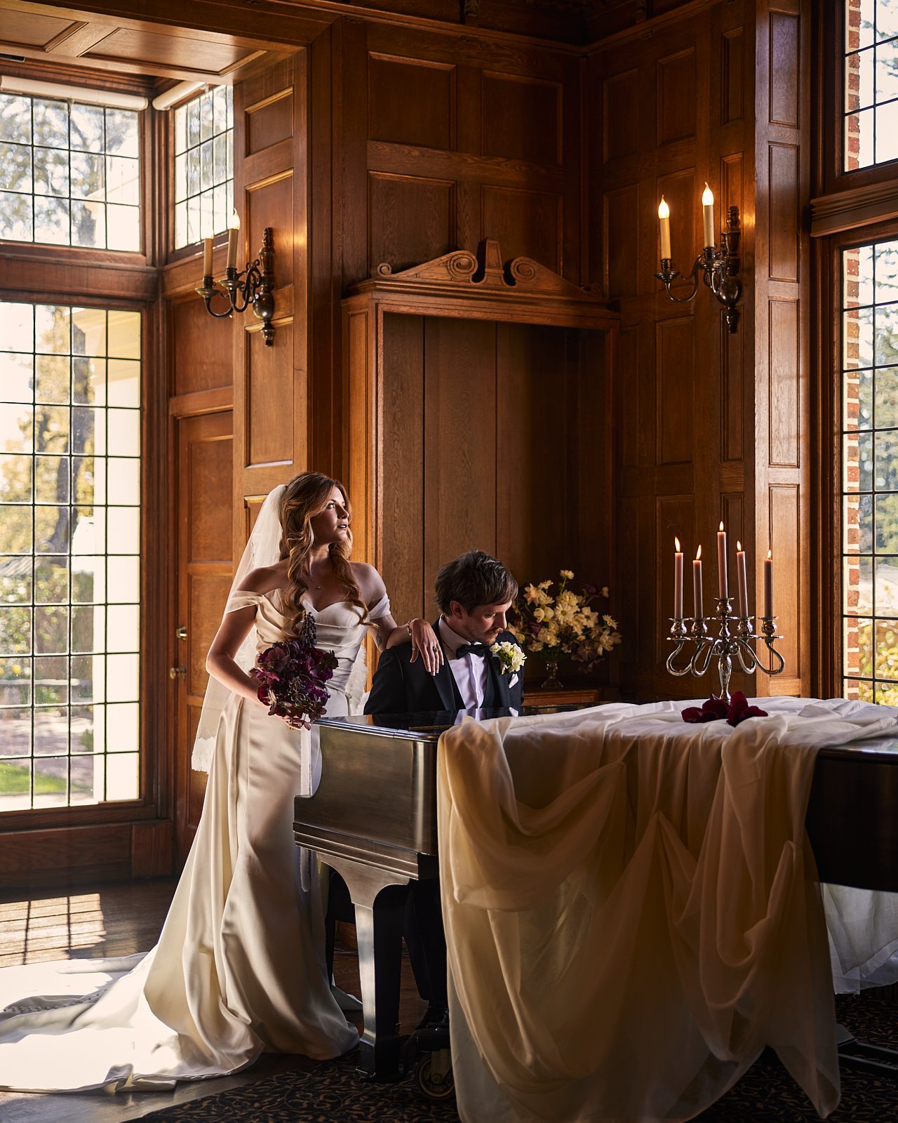 A bride and groom in a warmly lit, wood-paneled room during their wedding ceremony, with the bride standing next to the groom at a grand piano, holding a bouquet, and looking thoughtfully out the window.