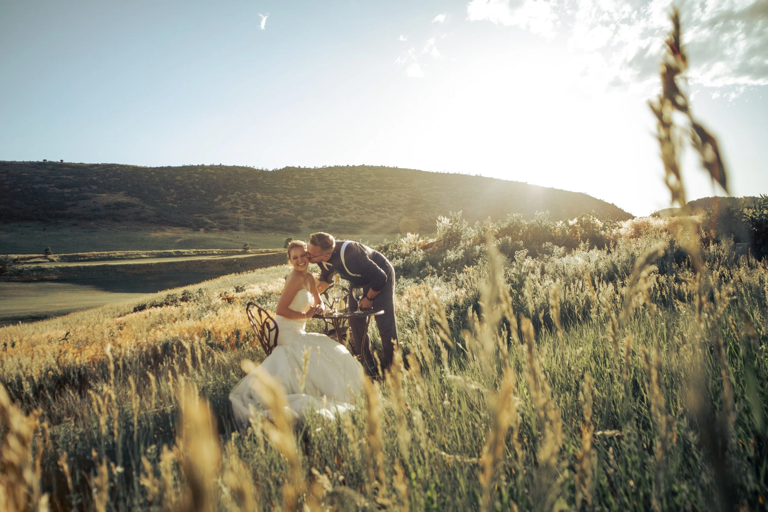 A bride and groom are having a romantic moment at a table set in a grassy field with wildflowers, during sunset with mountains in the background.