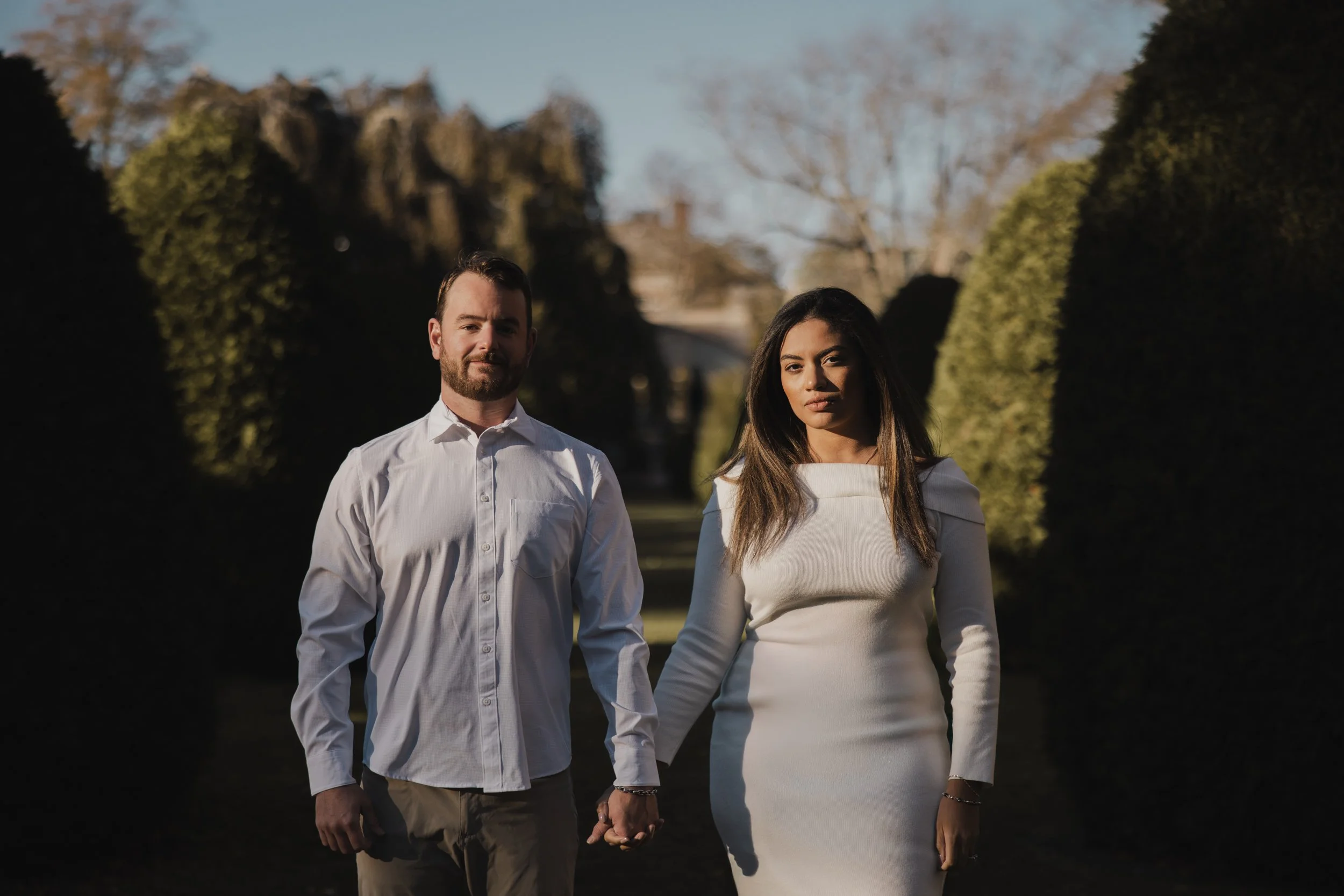 A man and a woman holding hands and walking outdoors during daytime, with shadowed greenery on either side and trees in the background.