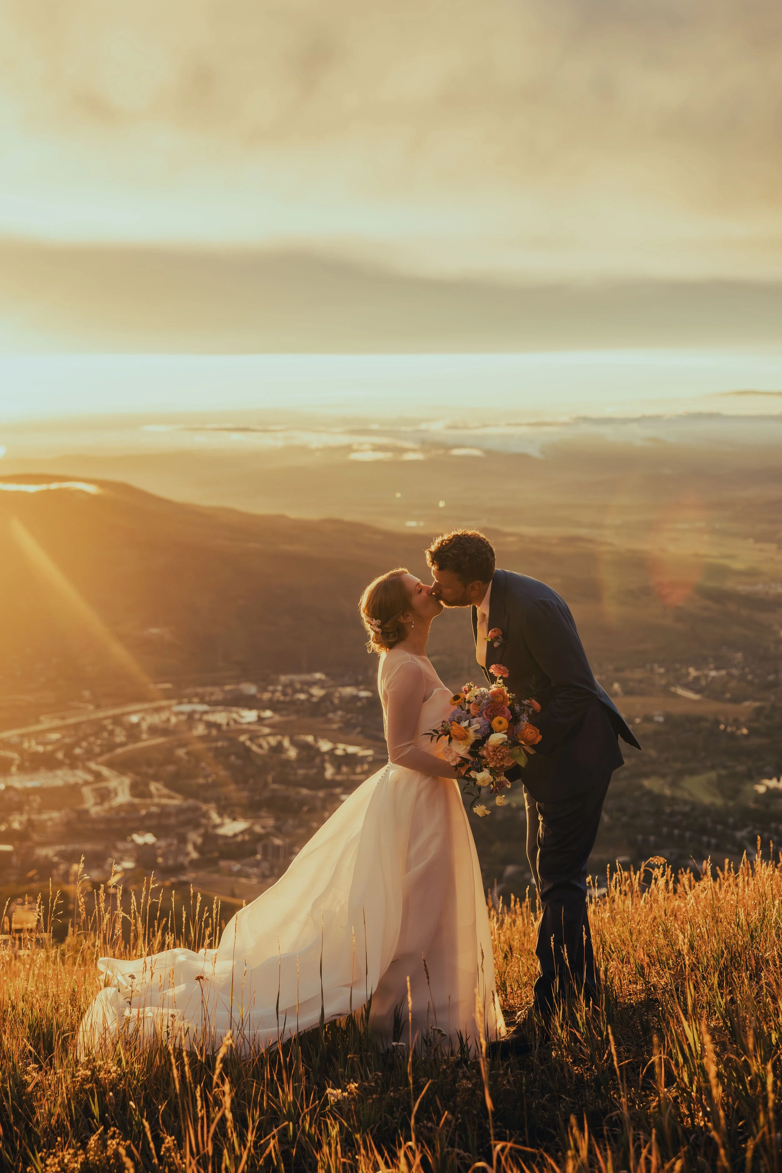 A bride and groom share a kiss during sunset on a hillside, with mountains and a valley in the background, and the bride holding a colorful bouquet.
