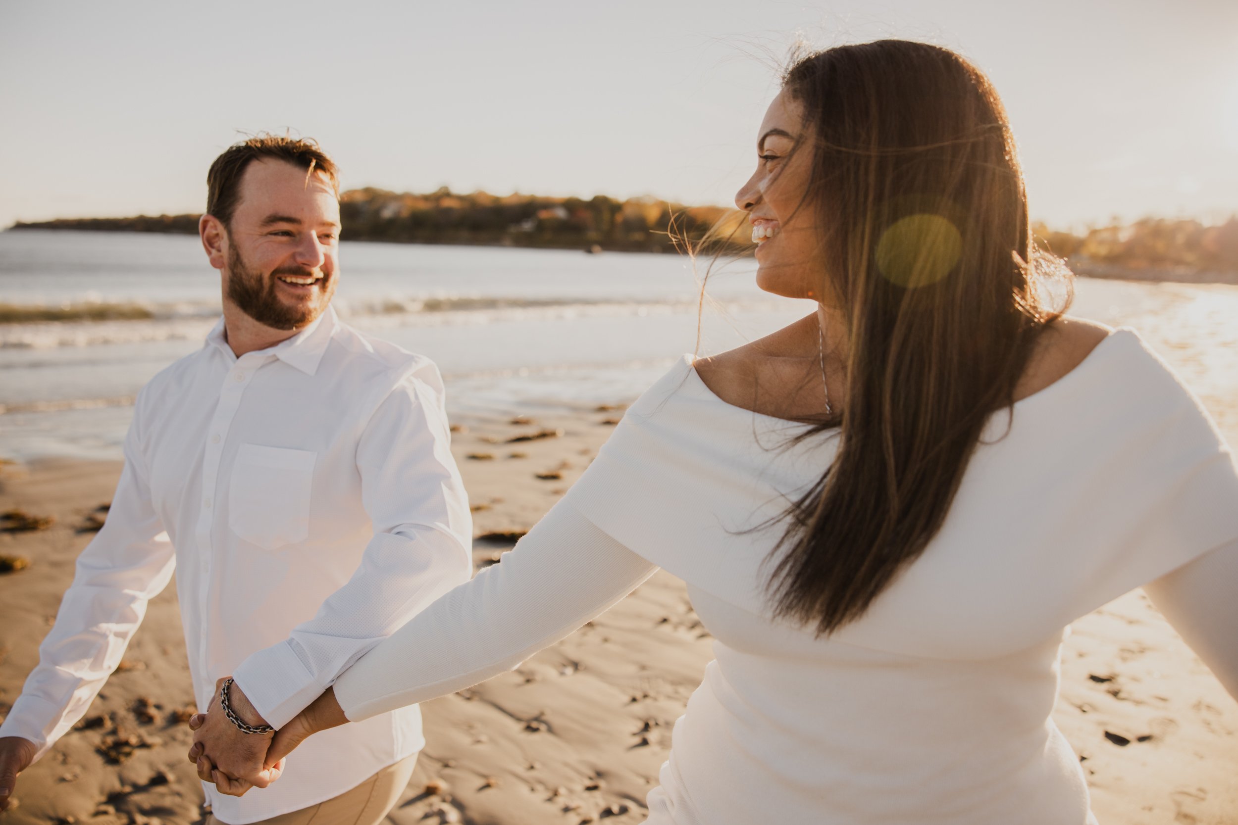 A smiling couple holding hands on a sandy beach during sunset, with waves in the background.