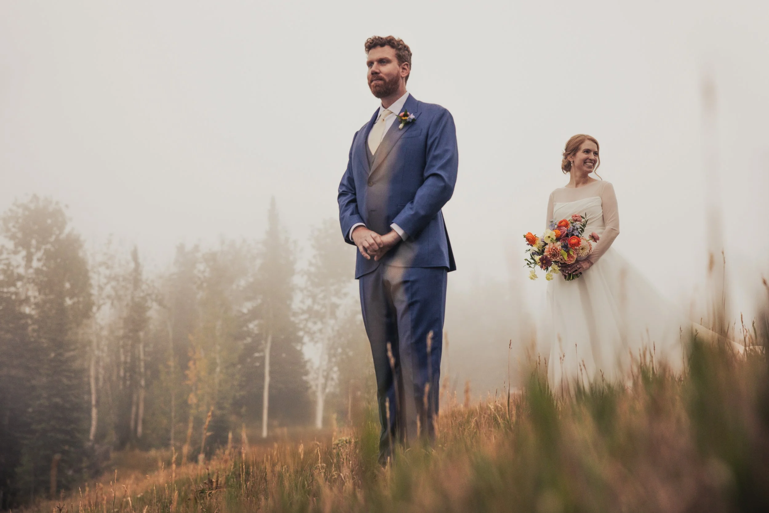 A groom in a blue suit stands with hands clasped, facing forward, with a bride in a white wedding dress holding a colorful bouquet, standing behind him in a misty outdoor setting with trees.