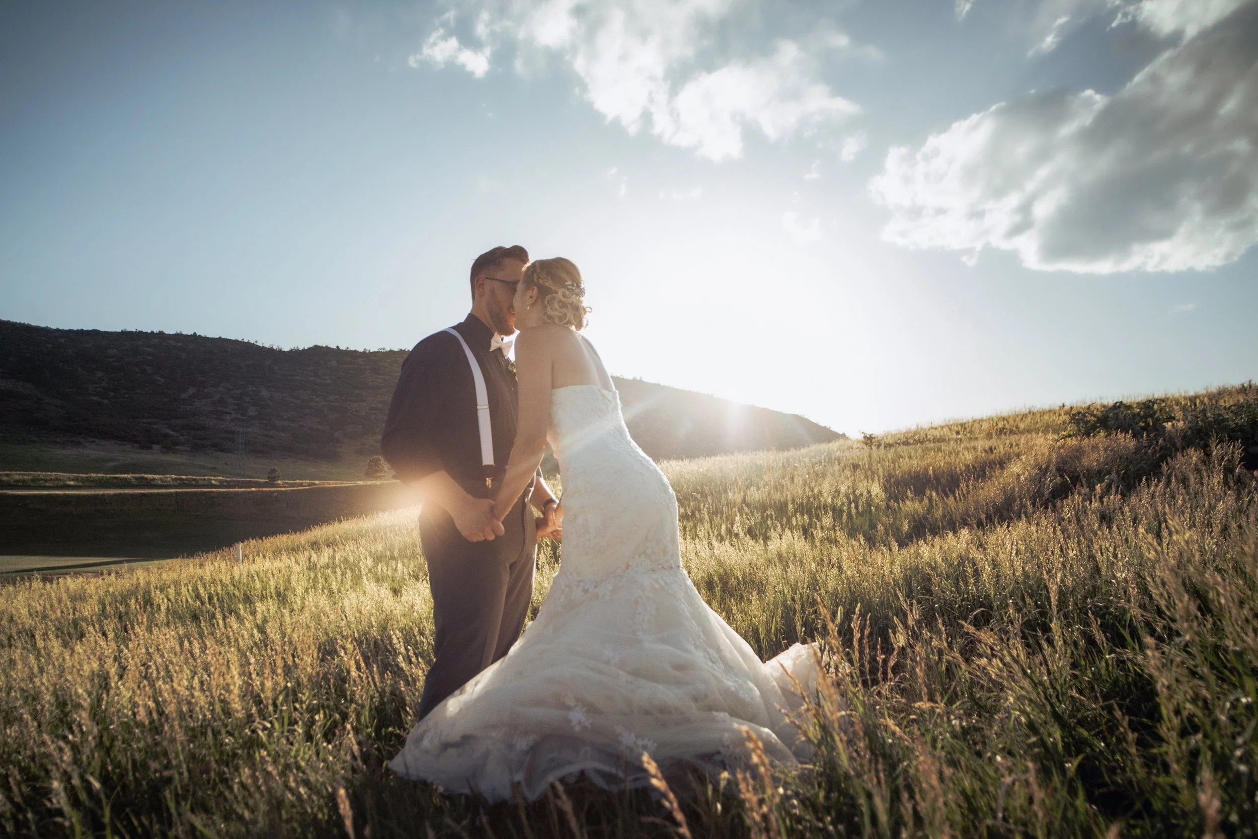 A bride and groom holding hands and kissing in a field with mountains in the background during sunset.