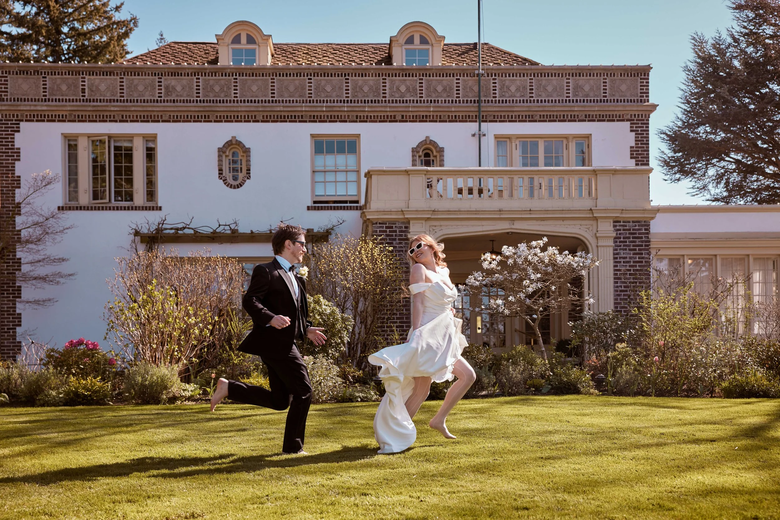 A bride in a white wedding dress and a groom in a black suit run barefoot on a green lawn in front of a large house with brick and white walls, windows, and a balcony. They are outdoors during daytime with trees and flowering bushes around them.