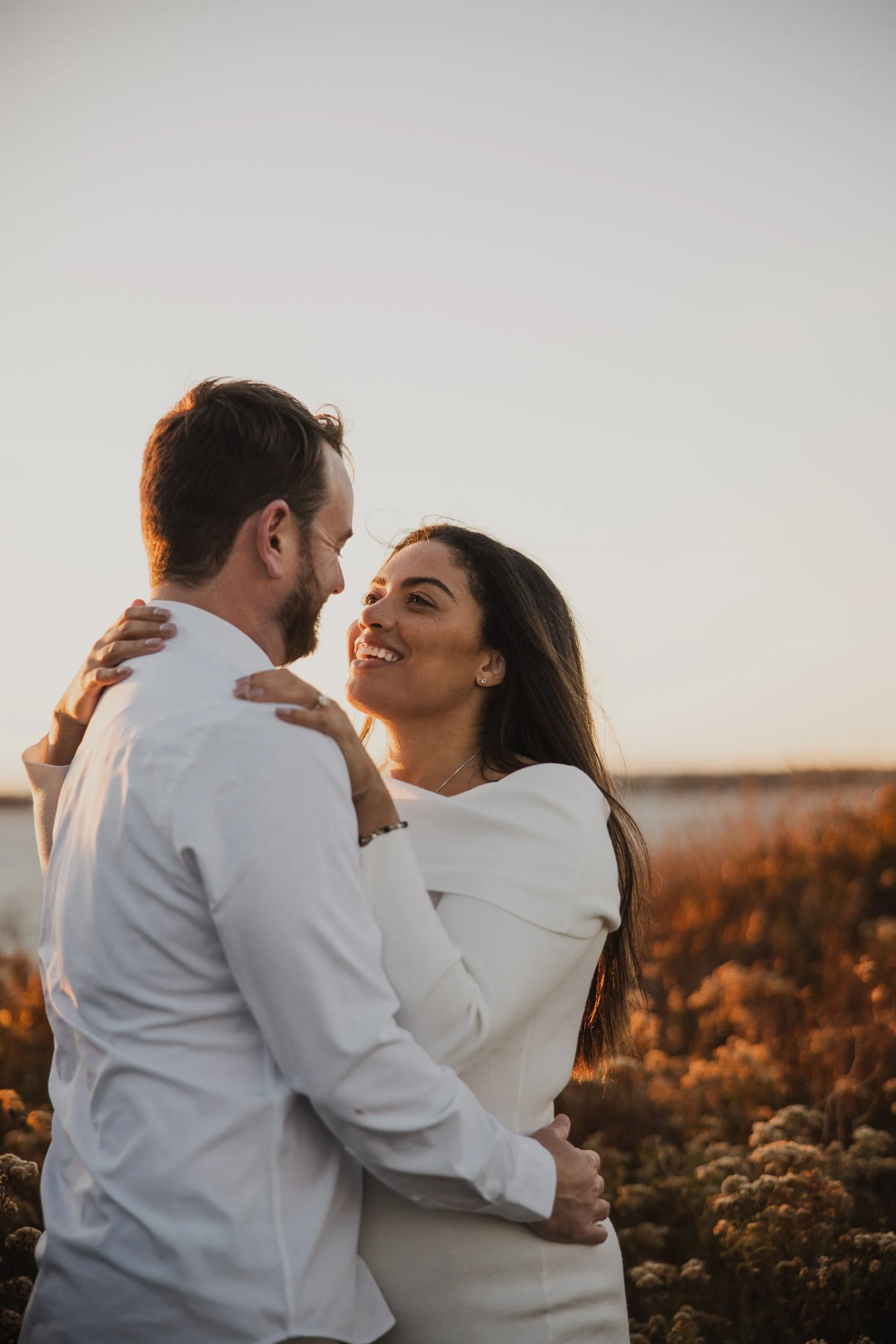 A couple happily embracing outdoors at sunset, facing each other and smiling, with a clear sky and natural landscape in the background.