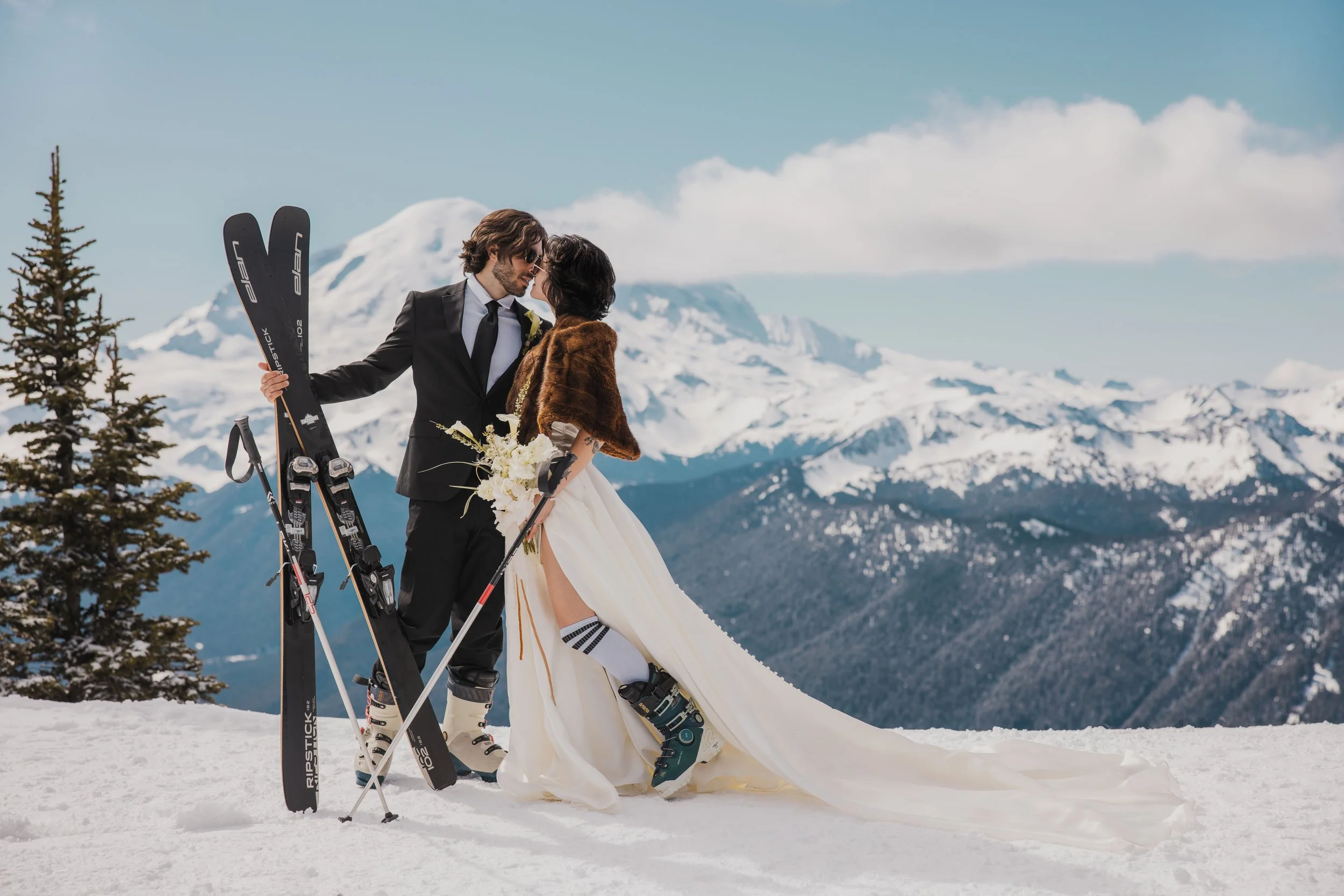 A newlywed couple stands in snowy mountains, dressed in wedding attire with ski equipment, sharing an intimate moment.
