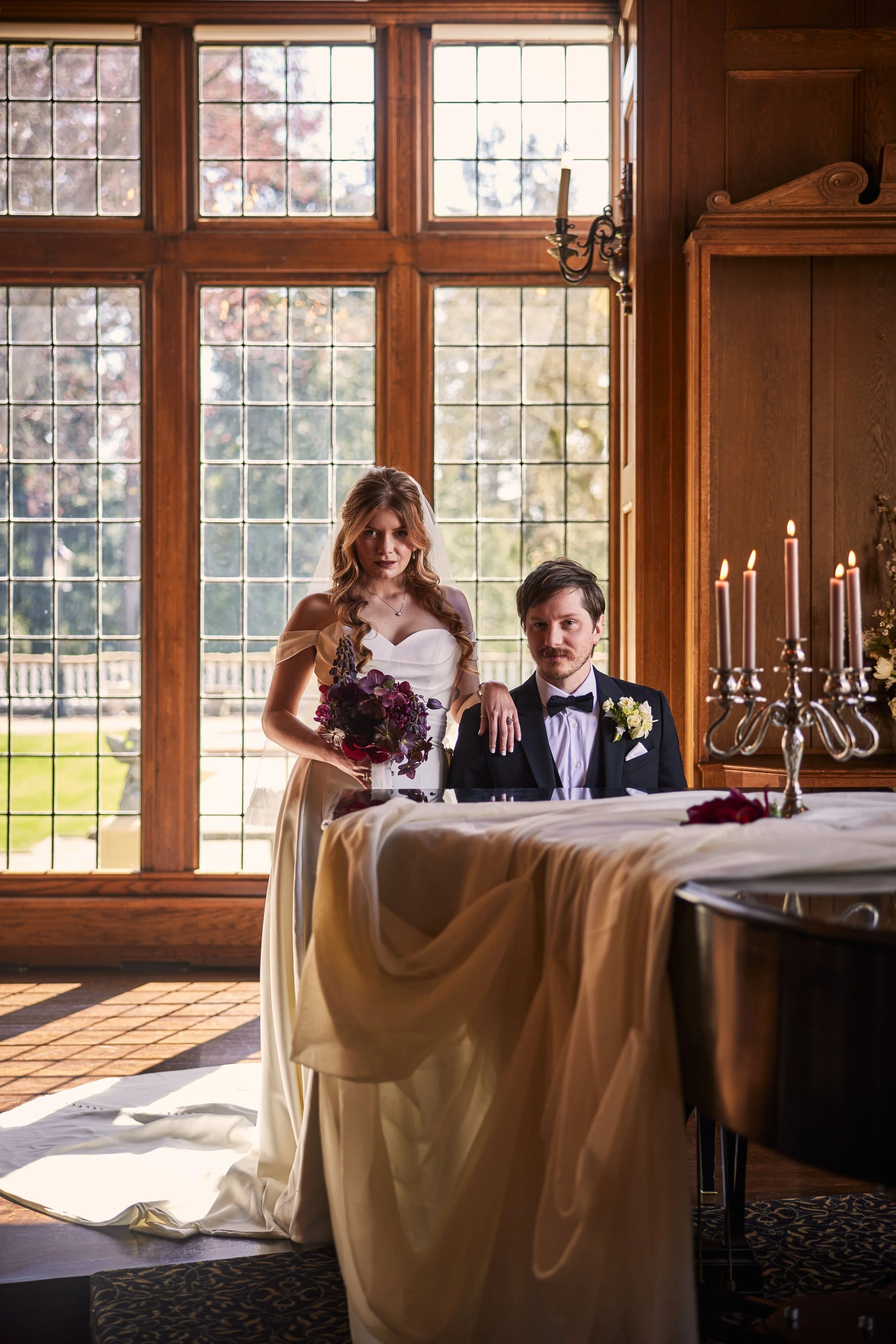 A bride and groom sit at a table in a room with large grid-patterned windows and wooden walls. The bride holds a bouquet of purple flowers, and the groom wears a tuxedo with a white boutonniere. There are lit candles on a candelabrum in front of them